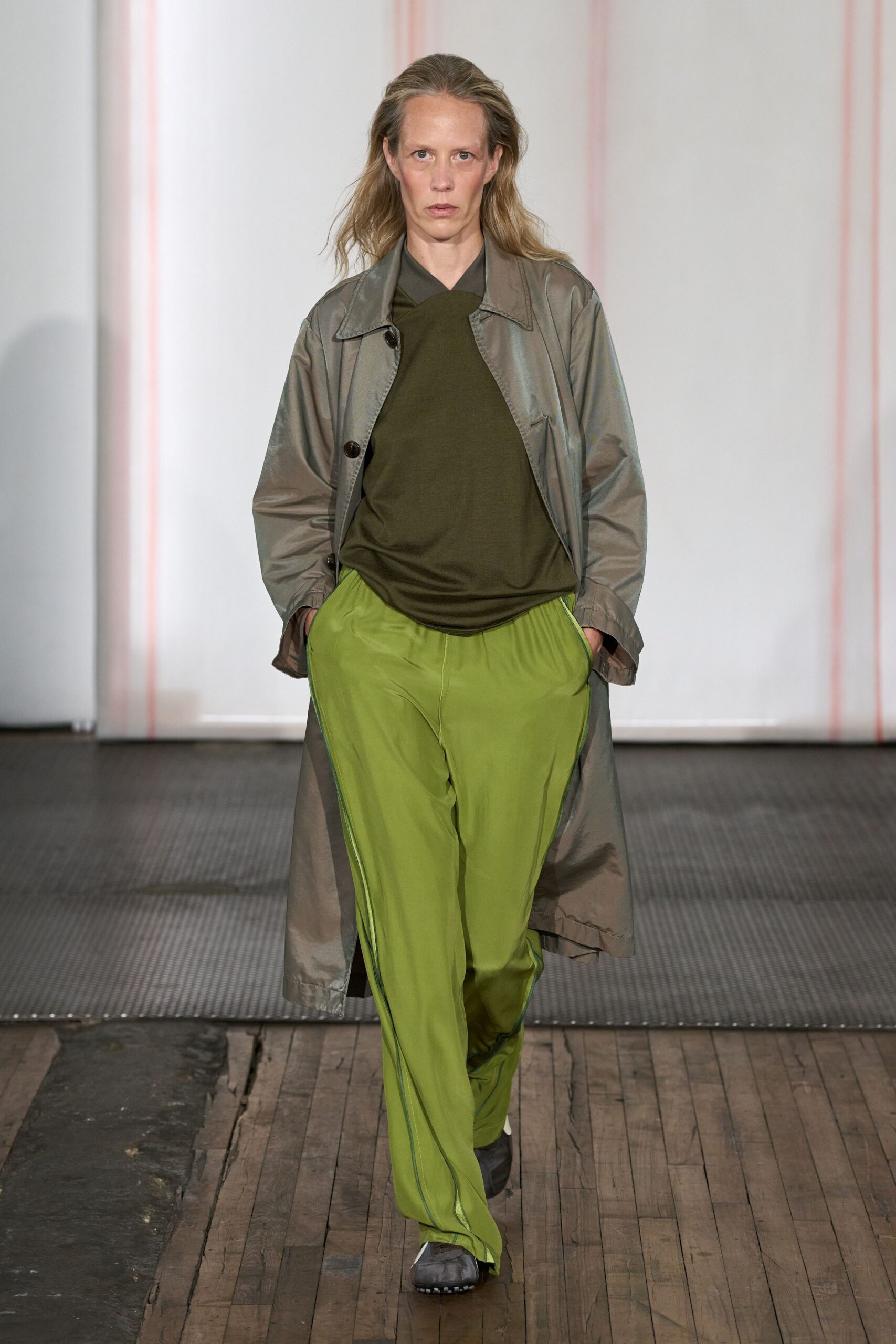 Taupe technical coat with a faint sheen and oversized dark buttons. Olive jersey top draped into an off-center folded neckline. Acid-lime track pants in fluid nylon, blue piping skimming the side seam. Grey trainers with black tread peeking under the hem. Walking to buy groceries just after rain, still carrying yesterday's mood.