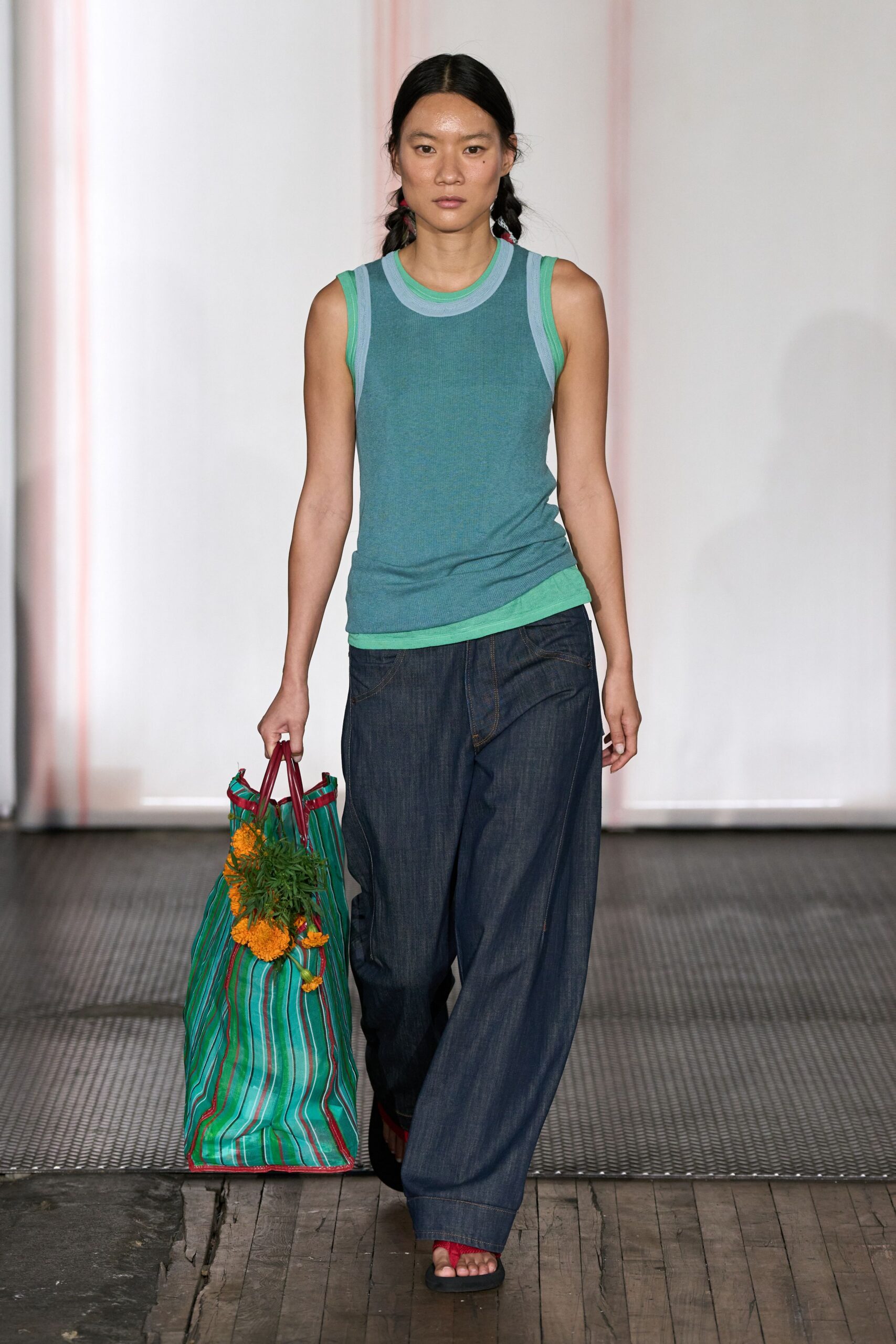 Blue-green ribbed tank layered over mint binding at neck and hem. Dark indigo wide-leg jeans with a low curved front seam. Striped market tote trimmed in burgundy, marigolds knotted to one side. Black flip-flops with bright red thong straps. Walking back from the flower market with stems brushing your calf.
