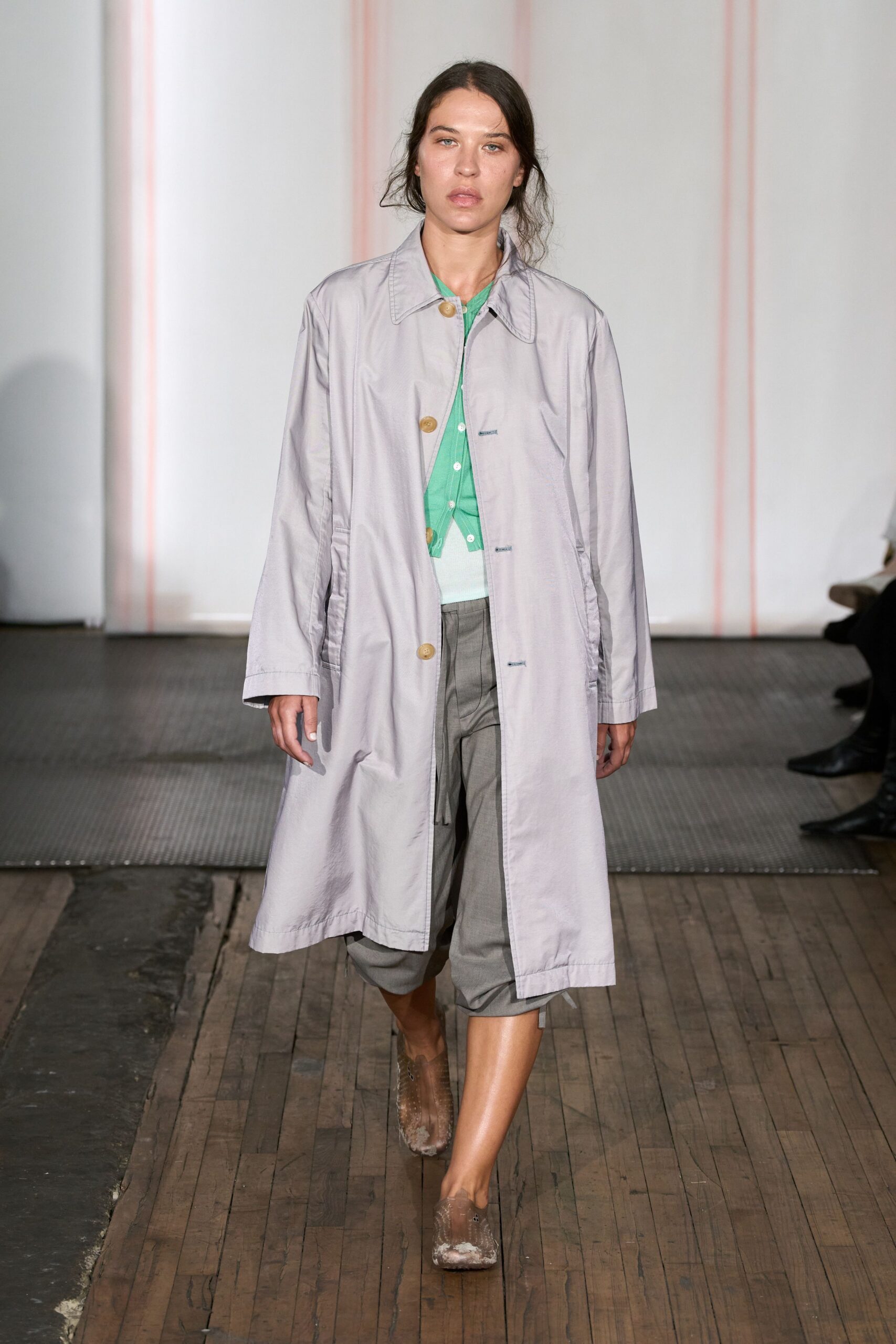 Pale lilac raincoat in crisp cotton, fastened with oversized horn buttons. Mint cardigan peeking through, a sliver of white knit underlayer. Grey cropped trousers with deep front pleats and a dropped crotch. Taupe mesh flats, toes dusted chalky white. Walking to buy groceries just after the pavement dries from rain.