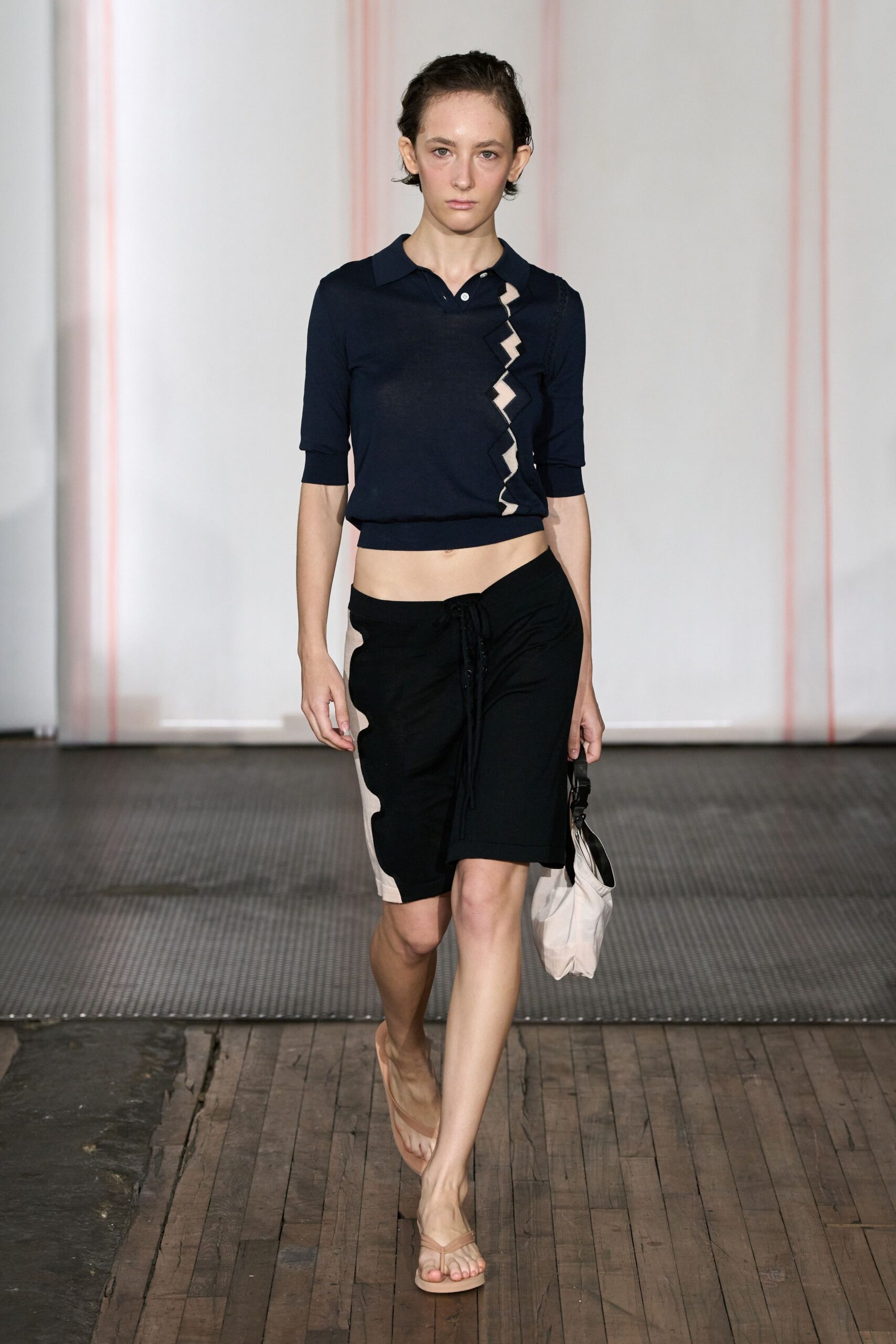 Navy fine-gauge polo knit with a cream zigzag cutout climbing one side. Black knee-length shorts with an off-centre drawstring and cream side inset. Slouchy chalk nylon bag with a black strap clip. Nude thong sandals, almost bare against the floor. Walking to buy iced coffee before the boardwalk heat turns sticky.