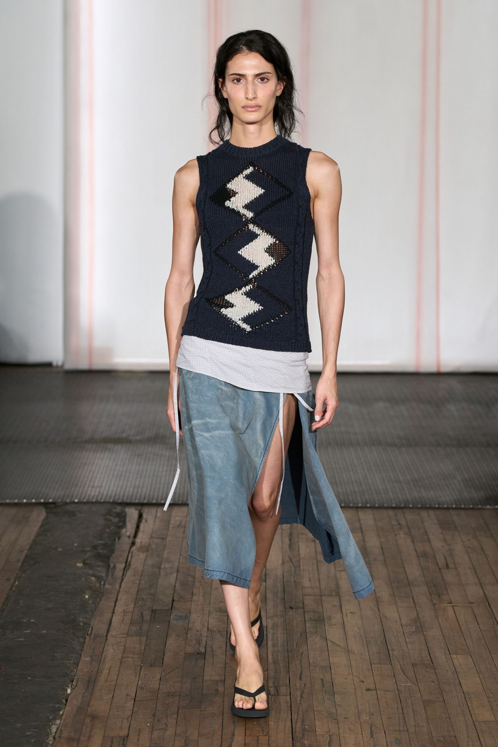 Navy open-knit shell with three cream intarsia diamonds down the front. Pale checked layer skewed low at the hip, ties hanging loose. Washed blue wrap skirt split high, one panel trailing past the calf. Black thong sandals. Walking the boardwalk before the sun burns off the sea haze.