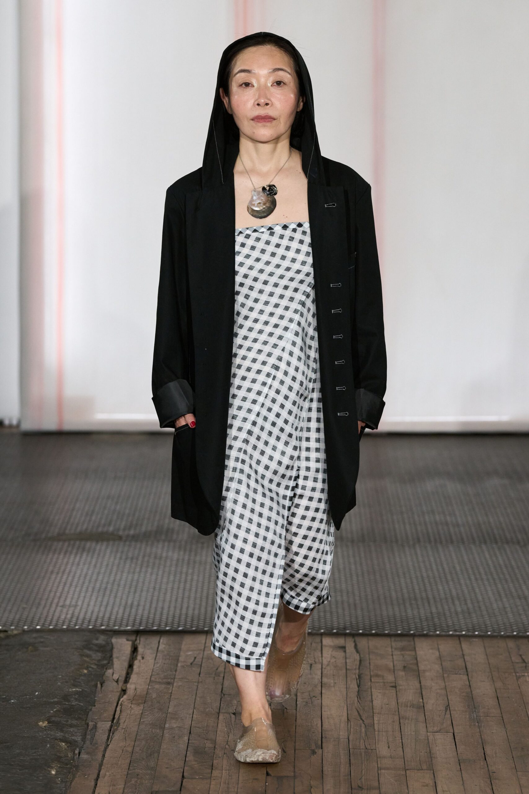 Black open coat with a built-in hood and rolled satin cuffs. Bias-cut strapless dress in black-and-white gingham, folding softly at the hip. Large shell pendant on a fine chain. Transparent mesh flats with cloudy marbled toes. Walking the boardwalk after rain, carrying your shoes in your hand.