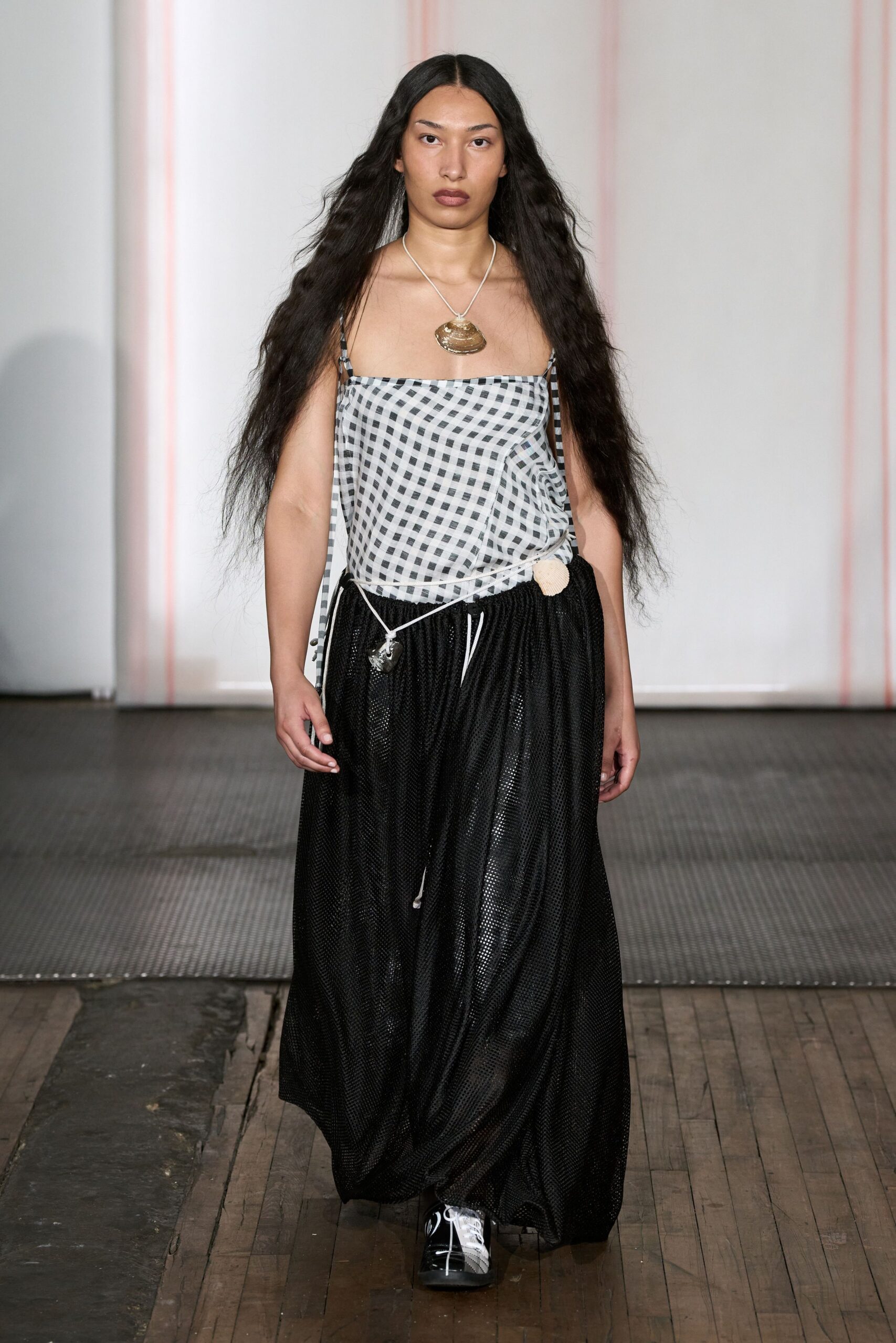 Black-and-white gingham camisole, cut on the bias with trailing side ties. Large ridged shell pendant on white cord. Black open-mesh maxi skirt, gathered low with a shell tucked at the hip. Black leather lace-ups with white contrast facing. Walking back from the pier with sand still stuck to one ankle.