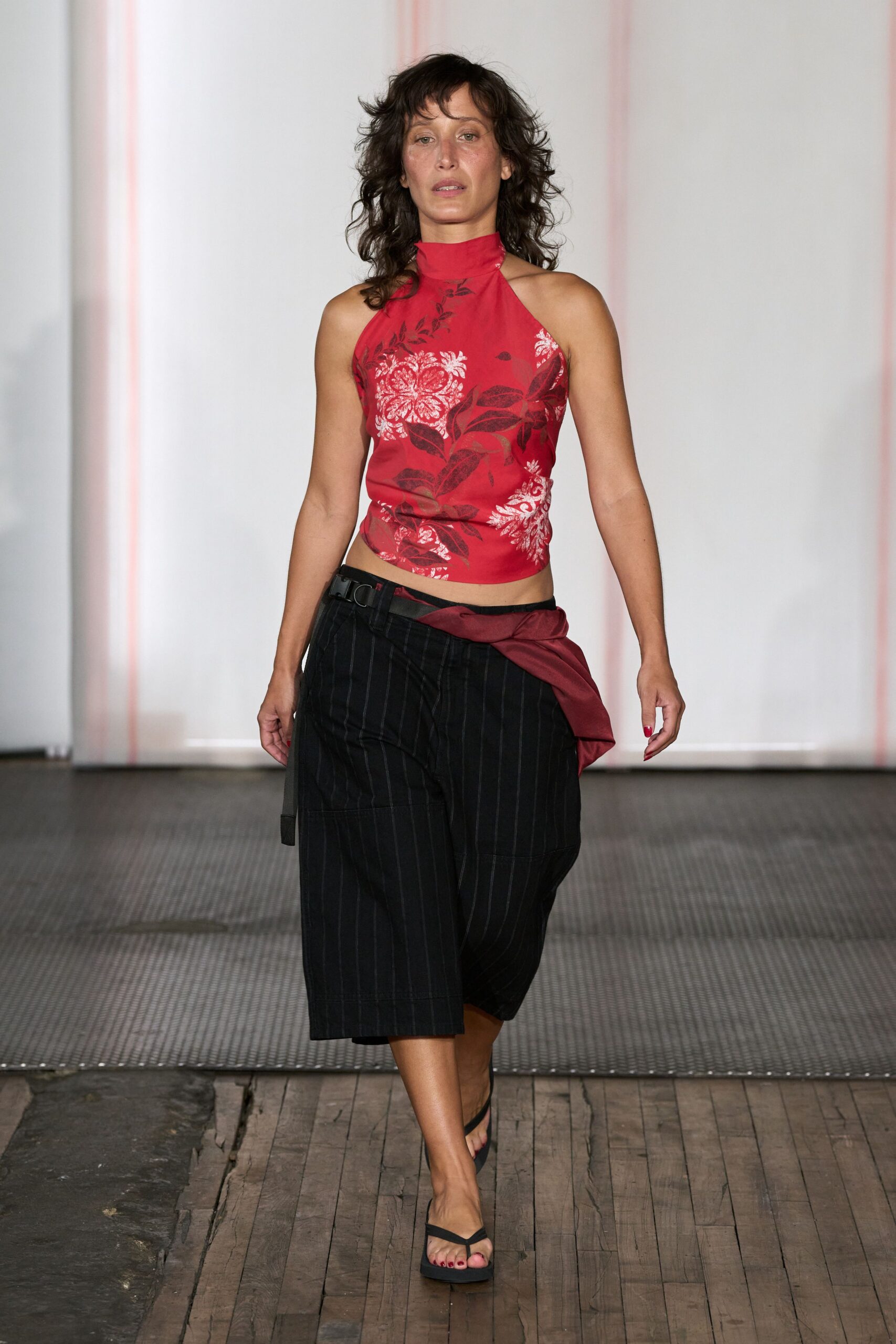 Cherry halter top with washed hibiscus print and a high stand collar. Charcoal pinstriped knee-length shorts cut broad through the leg. Burgundy sash knotted off-center under a black utility belt. Black thong sandals with flat foam soles. Walking the boardwalk after dinner, warm wind lifting the receipt from your hand.