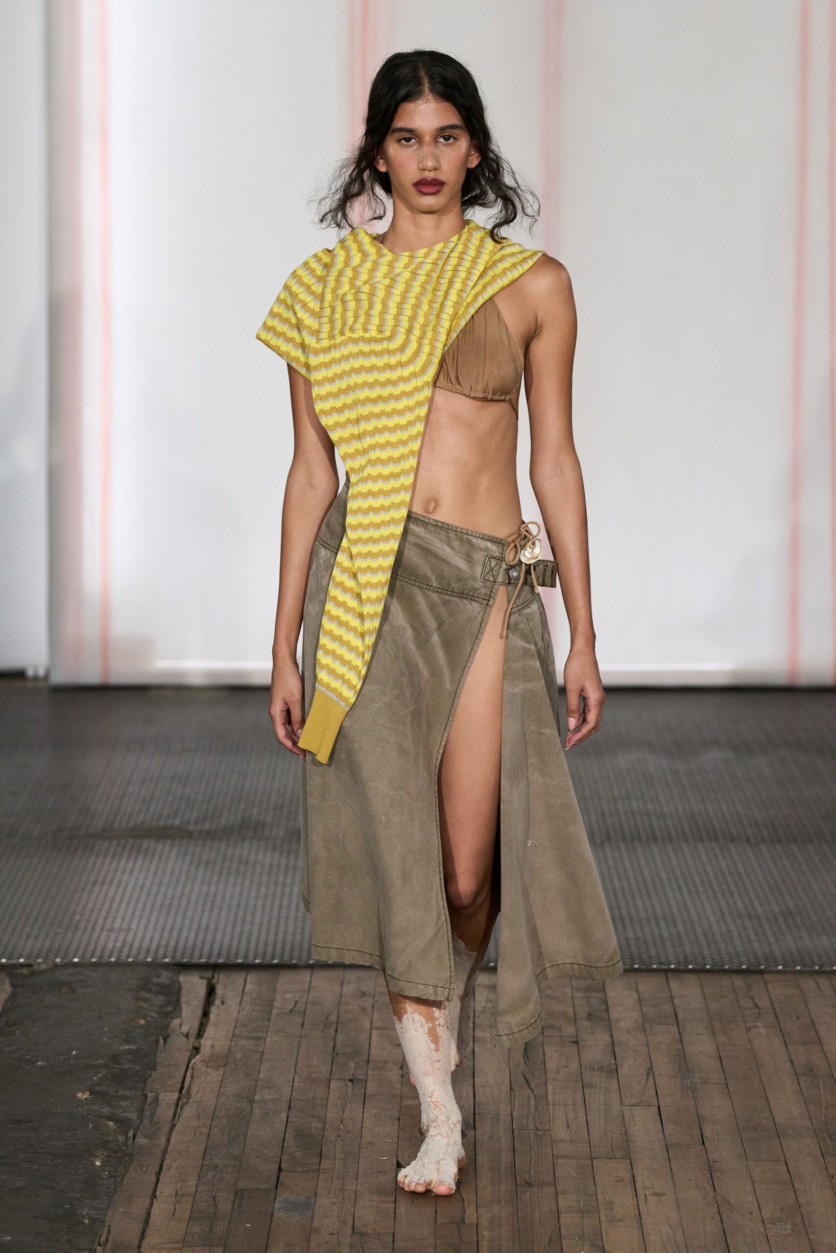 Yellow and white ribbed knit draped in a long off-centre panel. Taupe gathered triangle bralette, one cup left exposed. Washed khaki wrap skirt split to the hip, button tab at one side. White crusted coating caked from shin to bare toes. Crossing a tidal flat after the water has gone and not waiting for it back.