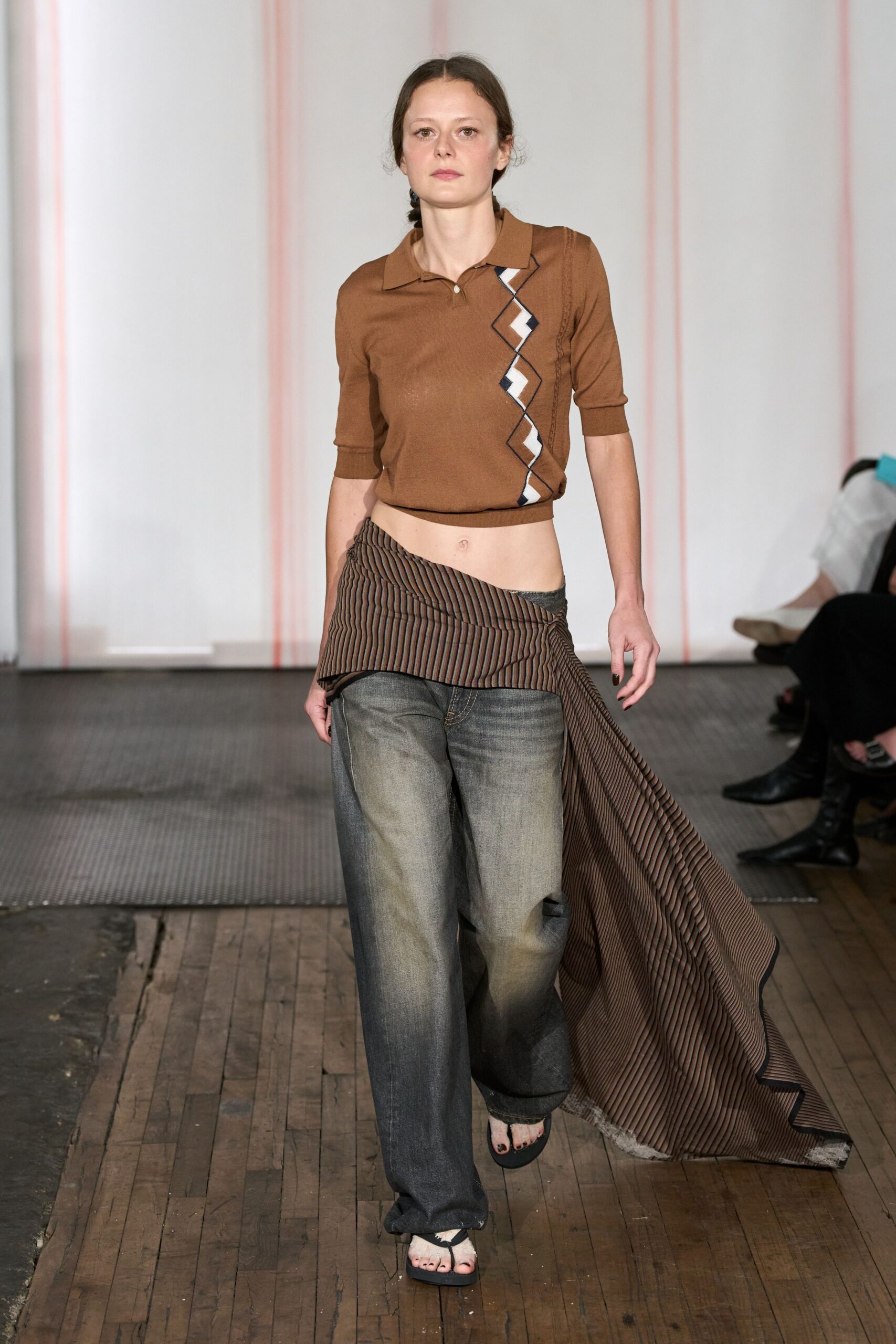 Cocoa fine-knit polo with a black-and-white zigzag intarsia panel. Brown pinstriped cloth knotted low, trailing in a long frayed sheet. Faded charcoal jeans cut loose through the leg. Black thong sandals with raw white fabric wrapped over the strap. Walking the boardwalk after swimming, denim hems darkening with salt.