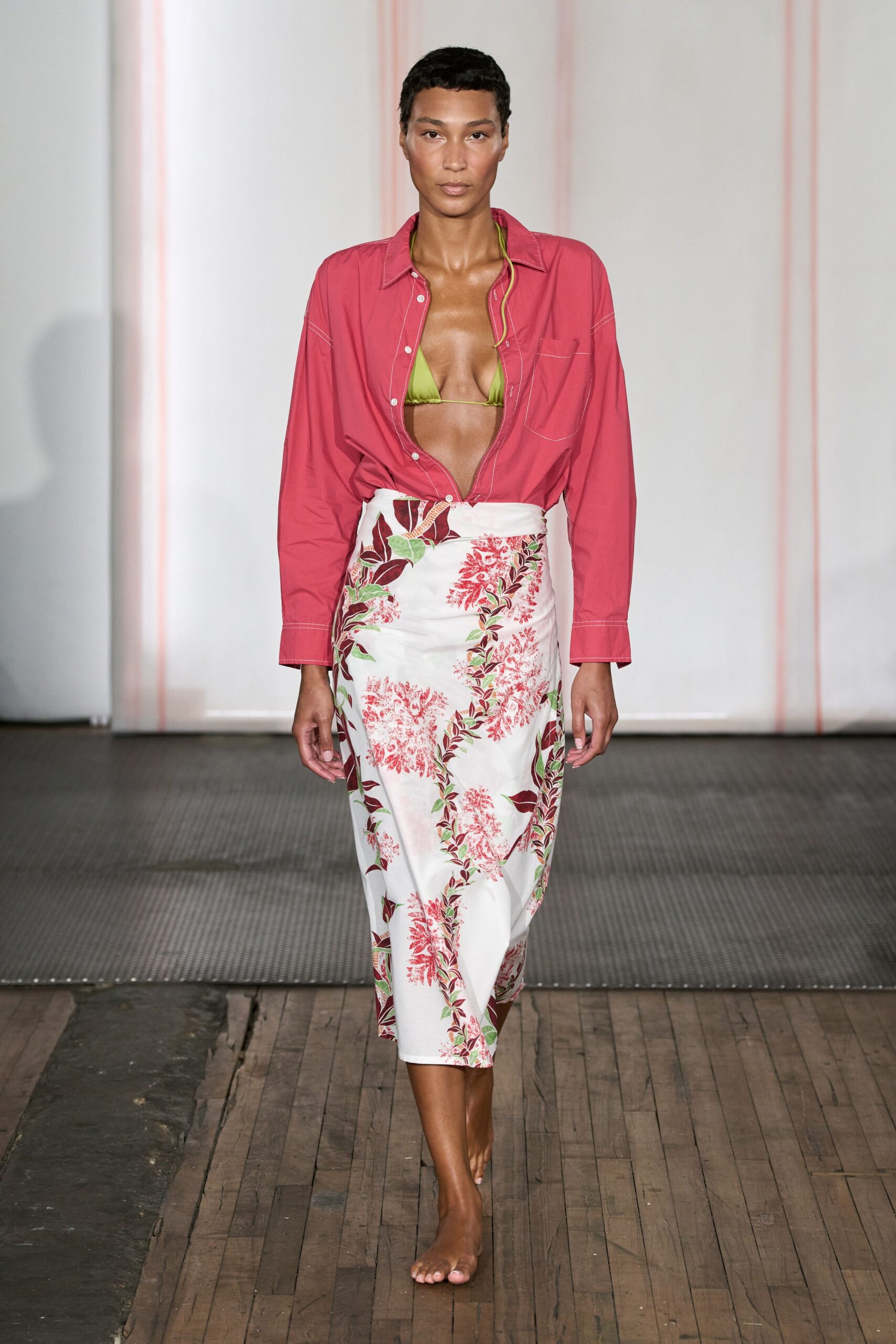 Watermelon cotton shirt hanging open, white topstitching tracing the pocket. Lime triangle bikini top on a thin string tie. White wrap skirt printed with coral sprays and brown leafy vines. No shoes, toes fully exposed. Walking back from the beach bar before the table is cleared.