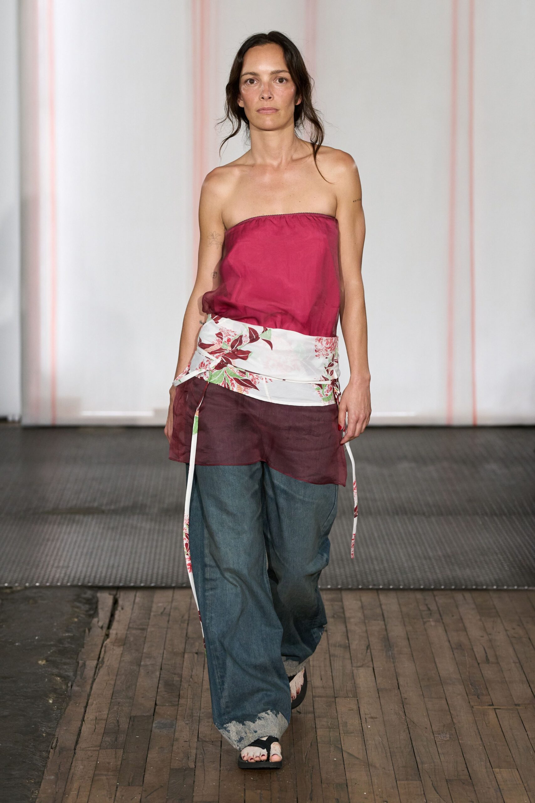 Sheer raspberry silk tube top with a softly gathered upper edge. White floral wrap tied low, two ribbon ends hanging past the thigh. Wide blue denim trousers with cloudy white abrasion around the hem. Black thong sandals with thick foam soles. Walking across sun-warmed boards after swimming, shirt still missing.