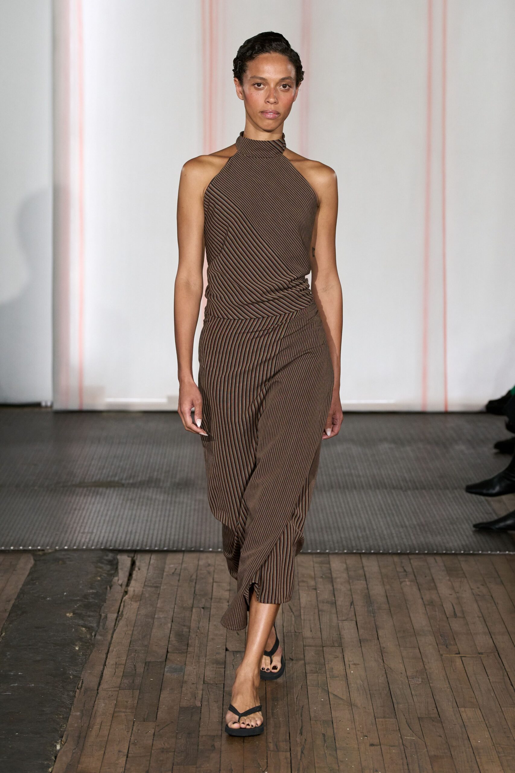 Brown micro-pleated halter, ribs running diagonally into a high neck. Matching wrap skirt with twisted drape and a narrow slit kicking forward. Black thong sandals with thick flat soles. Walking across sun-warmed boards after lunch, still tasting strong coffee.