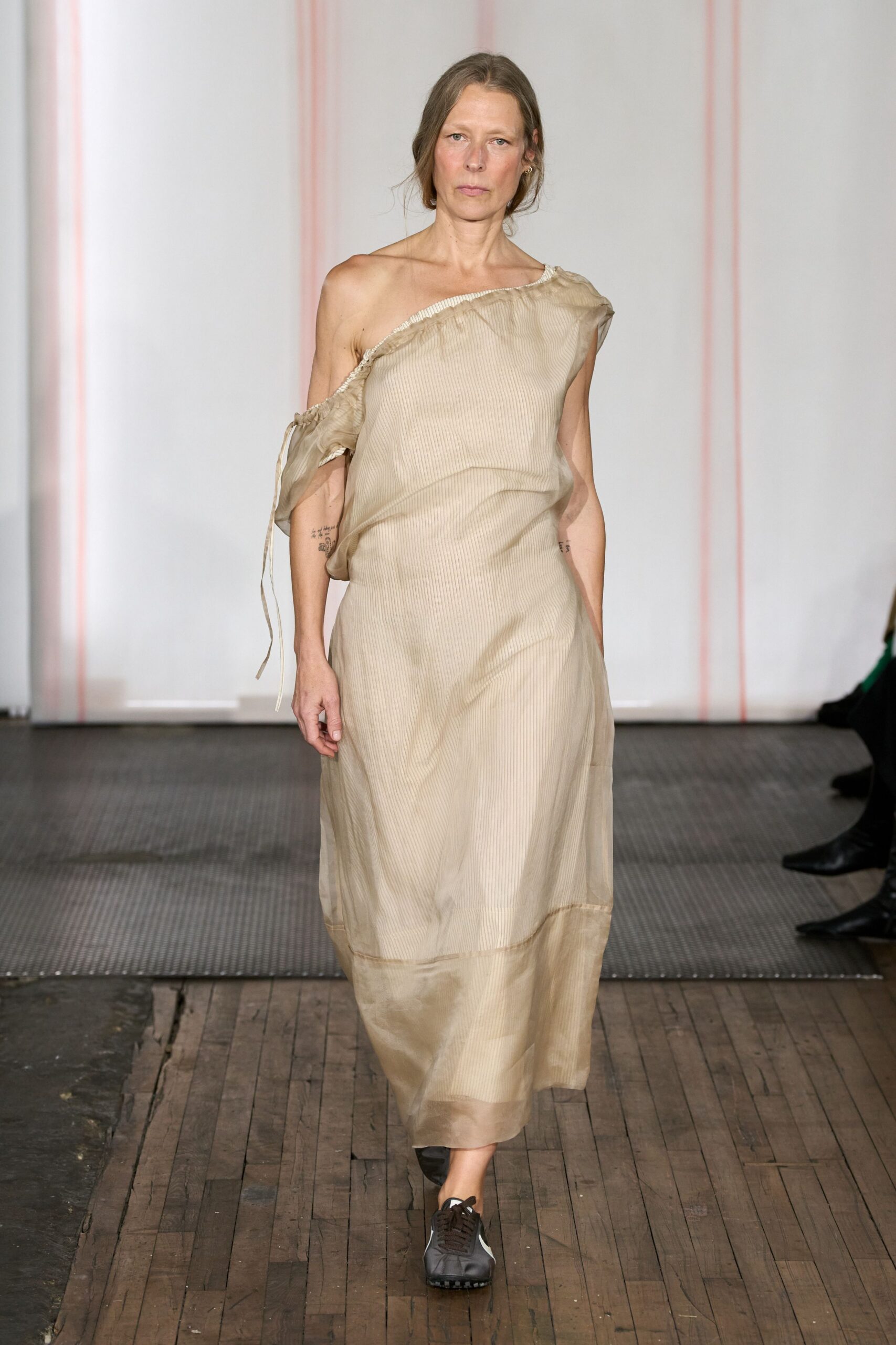 One-shoulder parchment dress in sheer pinstriped organza, raw ruffle edging and a dangling tie. Black track sneakers with white side panels and dark laces. Crossing a drafty town hall floor after changing out of dance clothes.