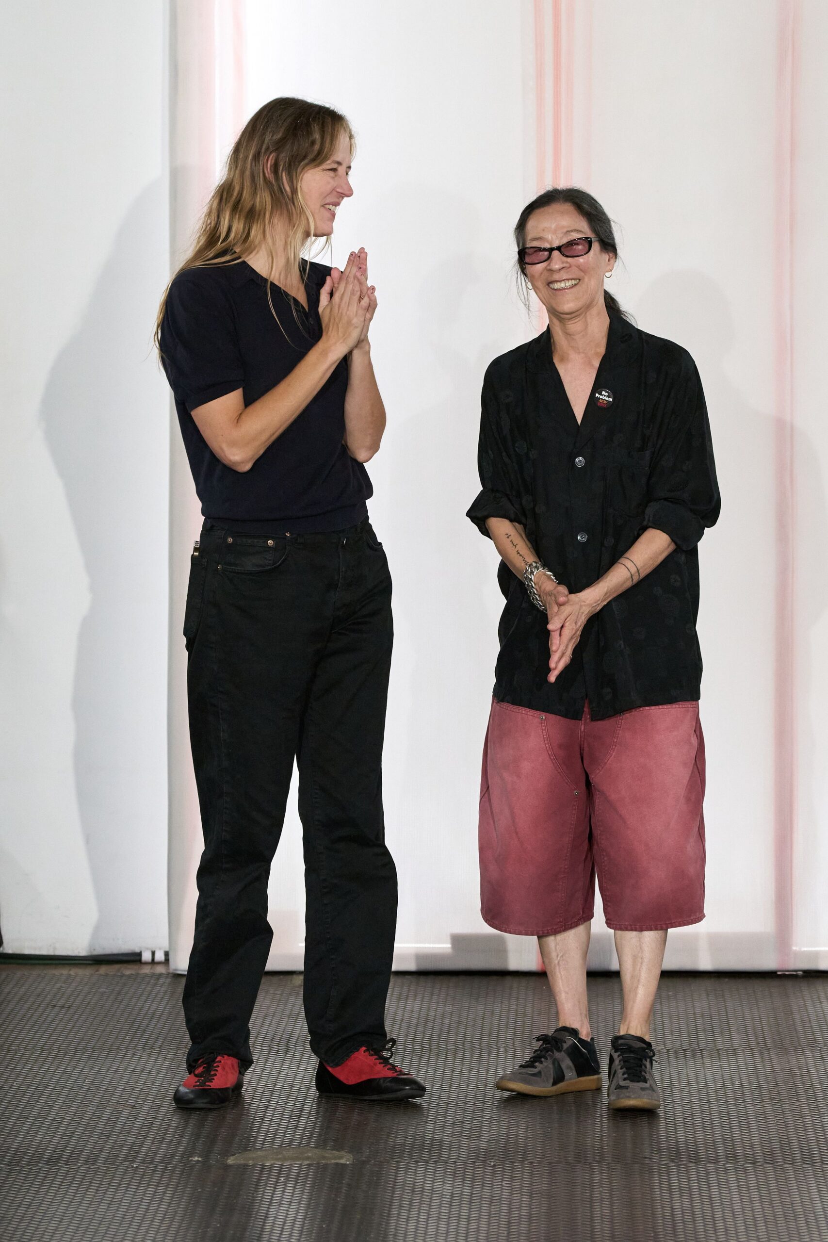 Narrow plum-tinted rectangular lenses. Black camp-collar shirt with rolled sleeves and shadowy tonal spots. Small round protest pin at the chest. Washed rose knee-length shorts with broad curved topstitching. Charcoal trainers with black side stripes and gum soles. After the talk, still smiling by the folding chairs.