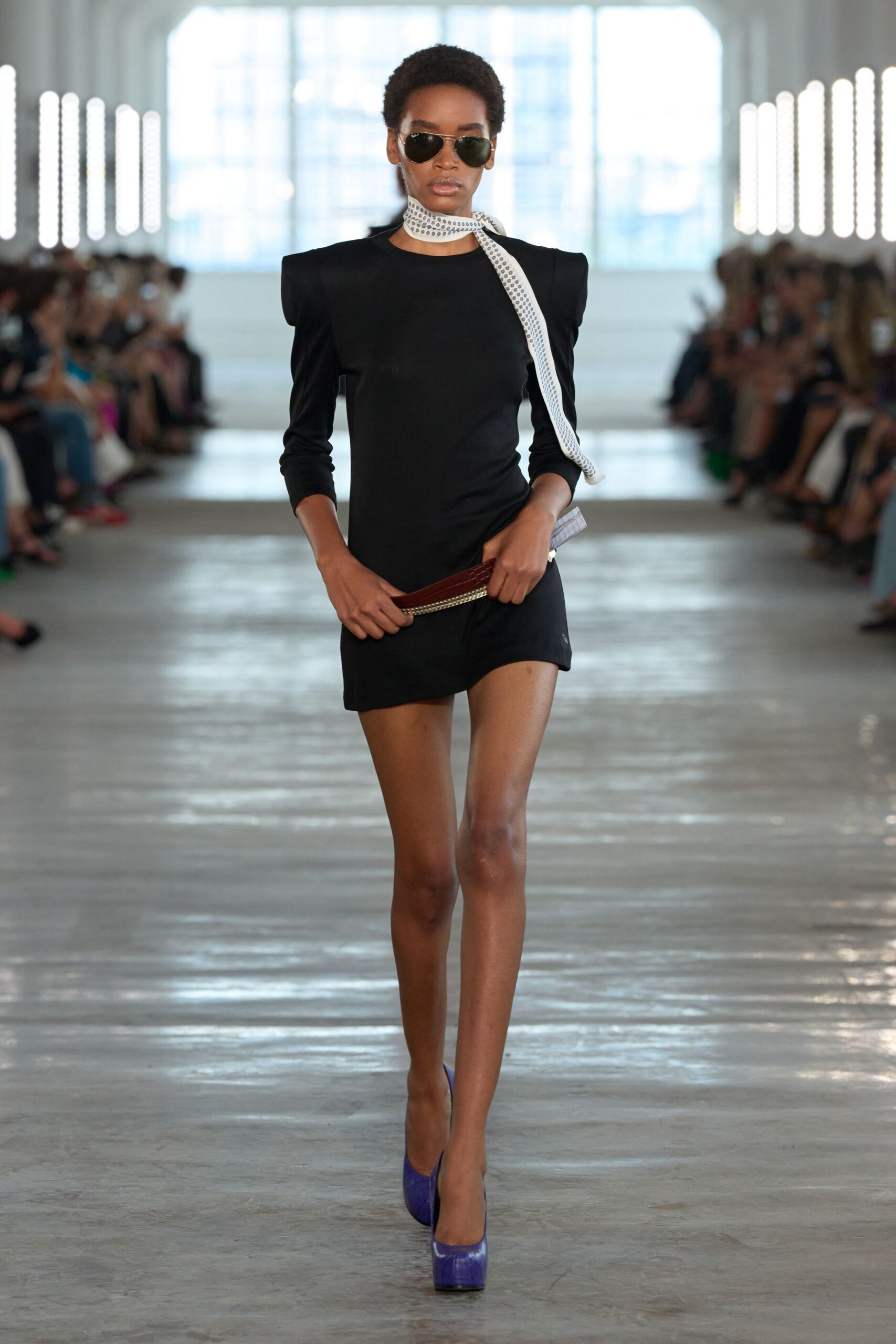 Dark aviators with a thin gold bridge. White eyelet scarf knotted close, one tail hanging long. Black jersey minidress with squared padded shoulders and a skewed hem. Burgundy croc-effect clutch with a rhinestone edge. Cobalt pumps in glossy leather. Crossing a hotel lobby before the wedding guests recognize you.