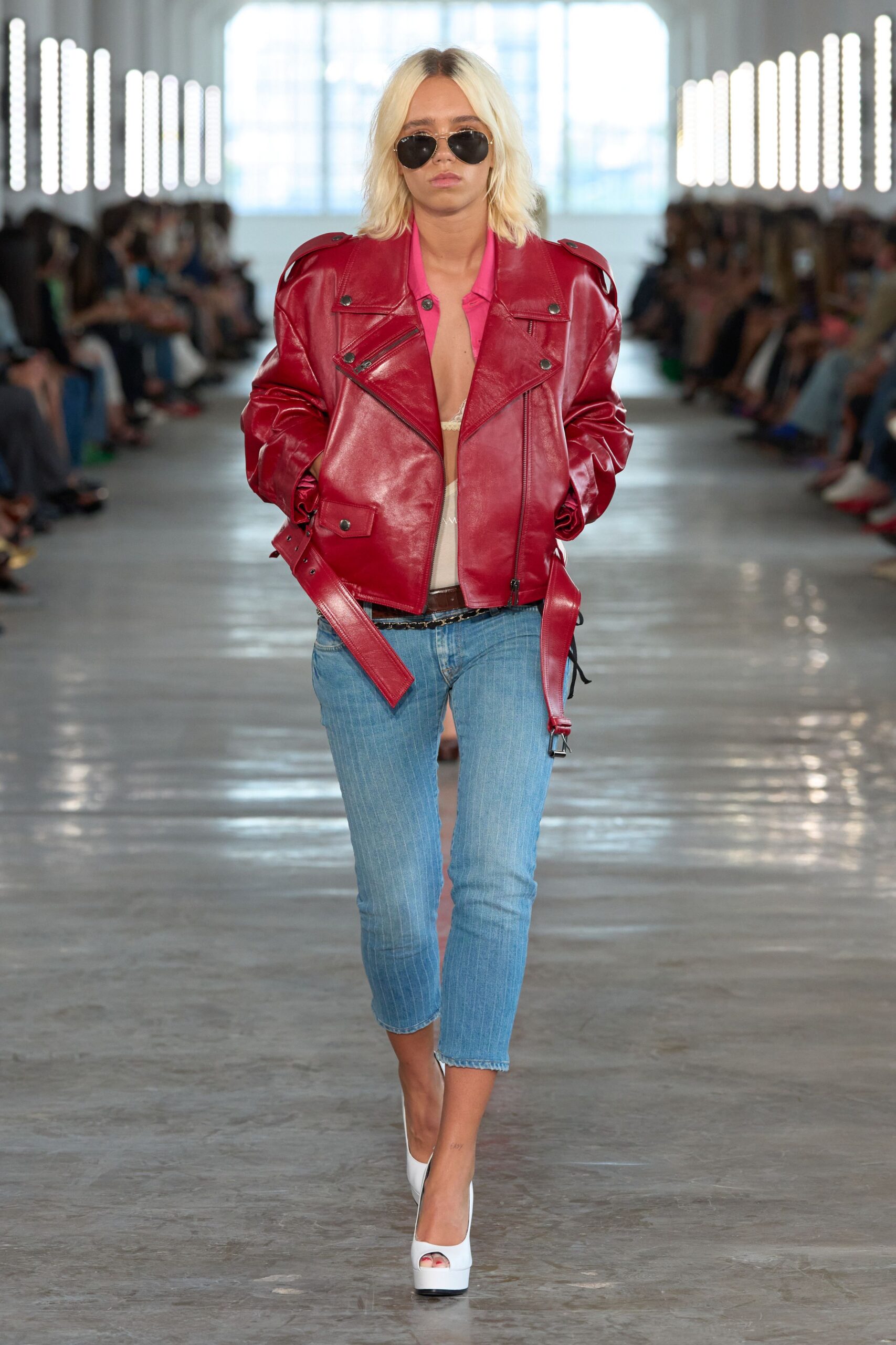 Black aviators with a thin gold bridge. Oversized cherry leather biker, belt strap swinging below the hem. Hot-pink shirt left open to the sternum. Cream lace triangle peeking at the center. Pale blue skinny jeans with a cropped raw hem. White peep-toe platforms. Walking out of a midday fitting into blinding summer pavement.