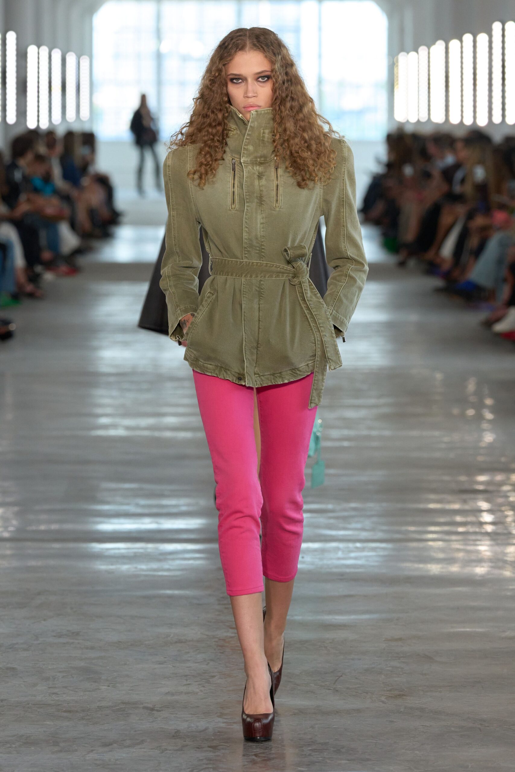 Olive twill jacket with a funnel neck and twin vertical chest zips. Self belt knotted off-center, one long end hanging past the hem. Hot-pink cropped leggings in dense stretch knit. Dark oxblood pumps with a thick platform sole. Walking to the corner shop after rain, still zipped against the wind.