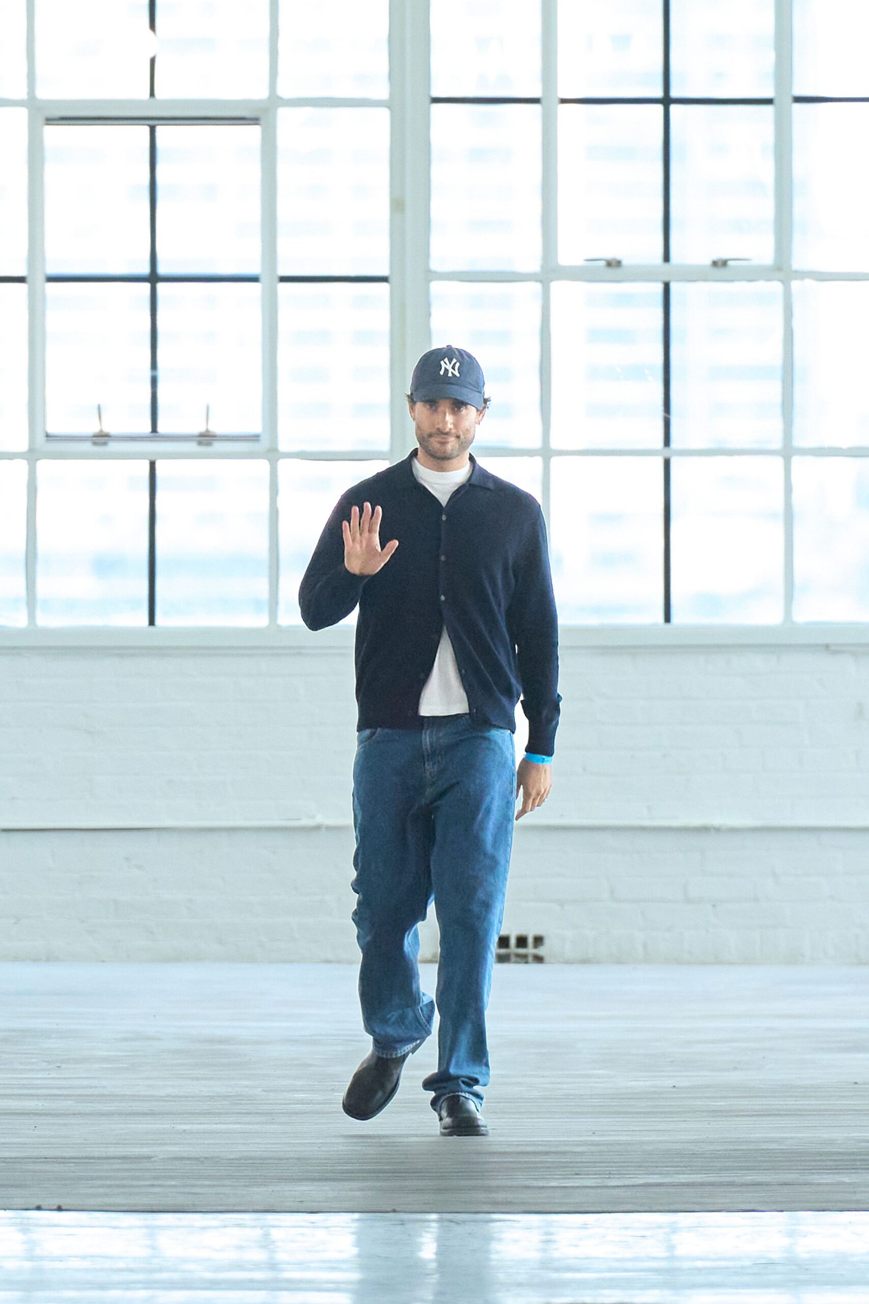 Navy twill cap with white front embroidery. Dark fine-knit cardigan, worn open with tiny tonal buttons. White crewneck tee peeking below the cardigan hem. Mid-blue straight jeans with a soft whiskered fade. Black leather slip-ons with rounded toes. Sunday studio cleanup, one window cracked against the heat.
