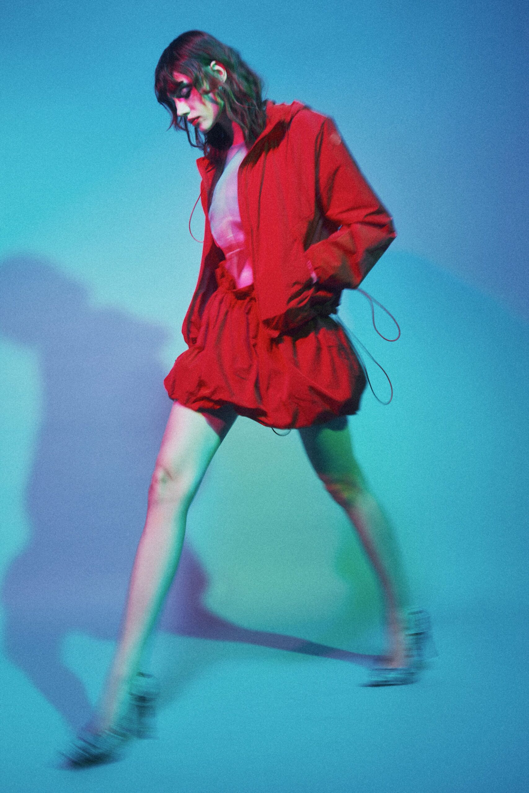 Crimson hooded jacket in crisp technical cloth, cords looping past the hips. Pale high-neck top with a smooth almost lacquered surface. Matching crimson bubble shorts, cinched waist and puffed hem. Black-and-white striped heels, blurred in motion. Crossing a color-washed studio floor before the fog machine clears.
