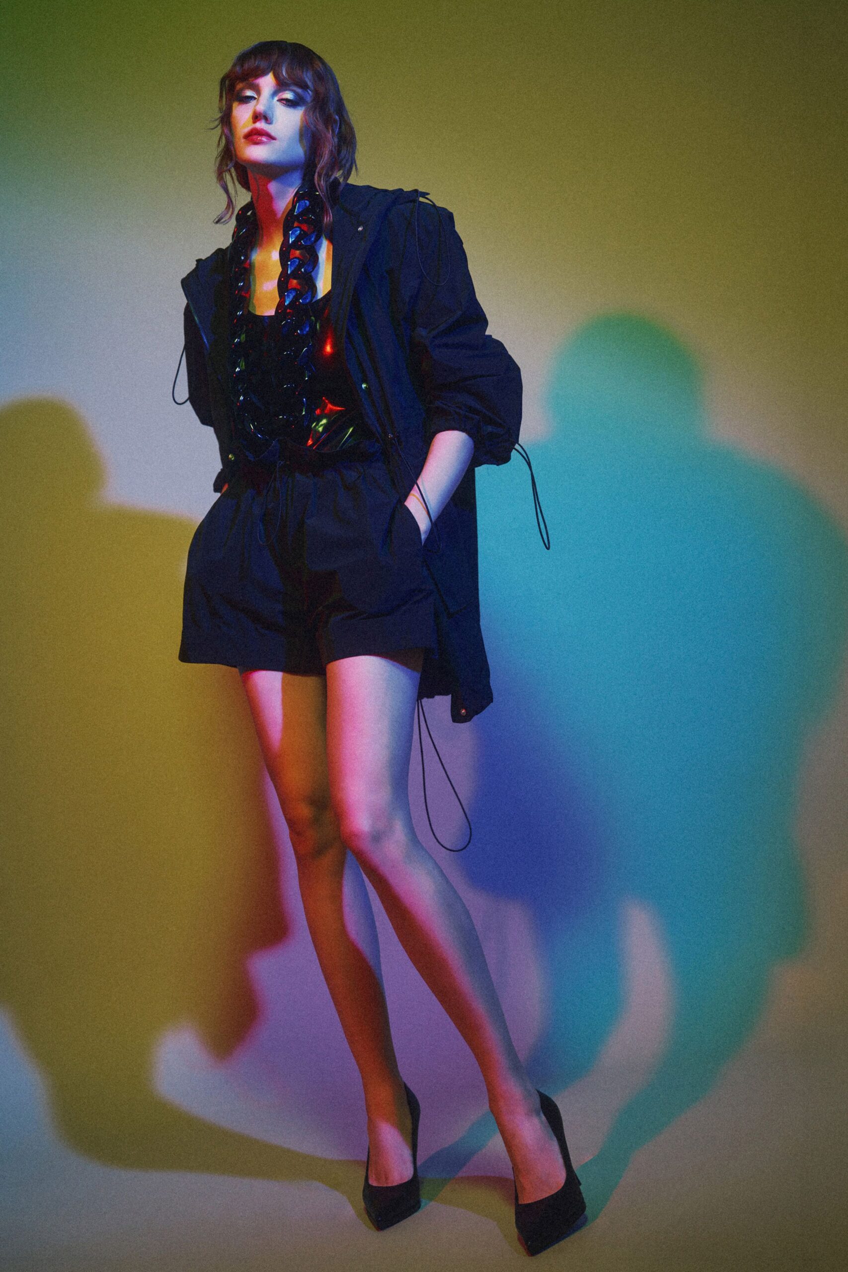 Black technical jacket with drawcords dangling past the hem. Black sequined camisole with a low square neckline. Oversized paillette scarf looped thickly around the neck. Paperbag shorts cinched tight with a self tie waist. Black pointed pumps. Waiting for the freight elevator after a party in an empty office tower.