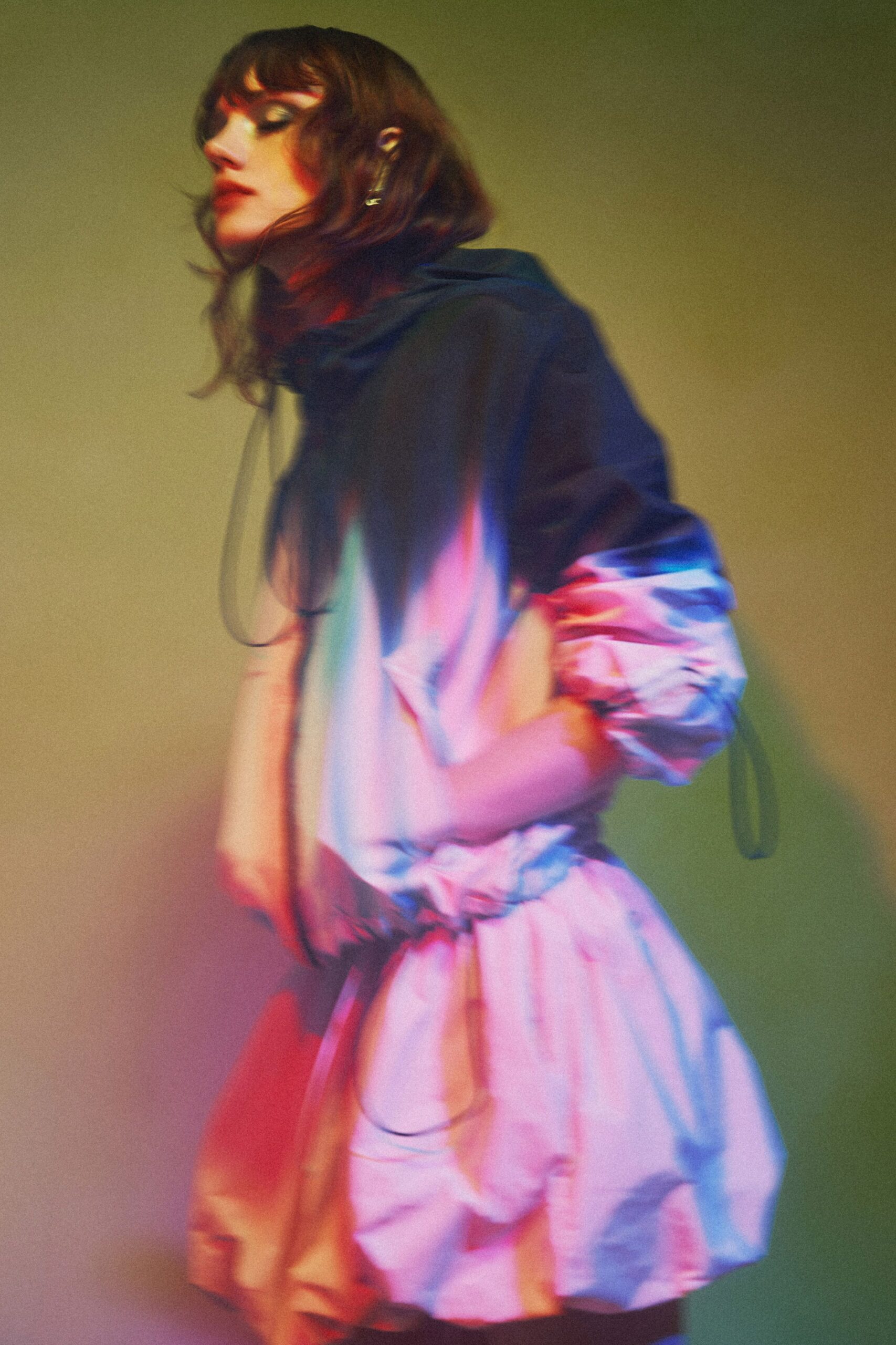 Iridescent windbreaker dress with a funnel hood and elastic-cinched waist. Puffed raglan sleeves gathered tight at the forearm with dangling cords. Crisp bubble hem flaring out in sculpted folds above the knee. Single slim drop earring. Crossing a motel parking lot while the neon sign keeps changing colour.