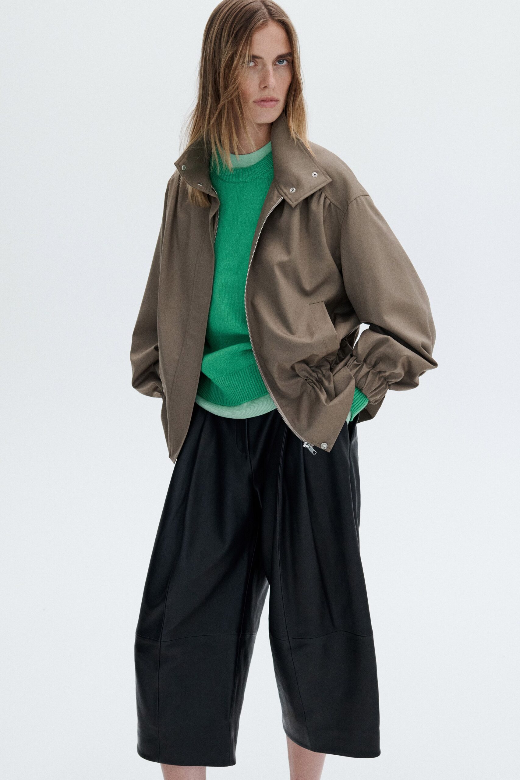Taupe cotton zip jacket with a snapped stand collar and gathered cuffs. Bright green fine-knit crewneck, mint underlayer peeking at neck and hem. Black leather culottes cut wide and long, center seam dropping straight. Walking to the corner shop just after rain, keys lost somewhere in the lining.
