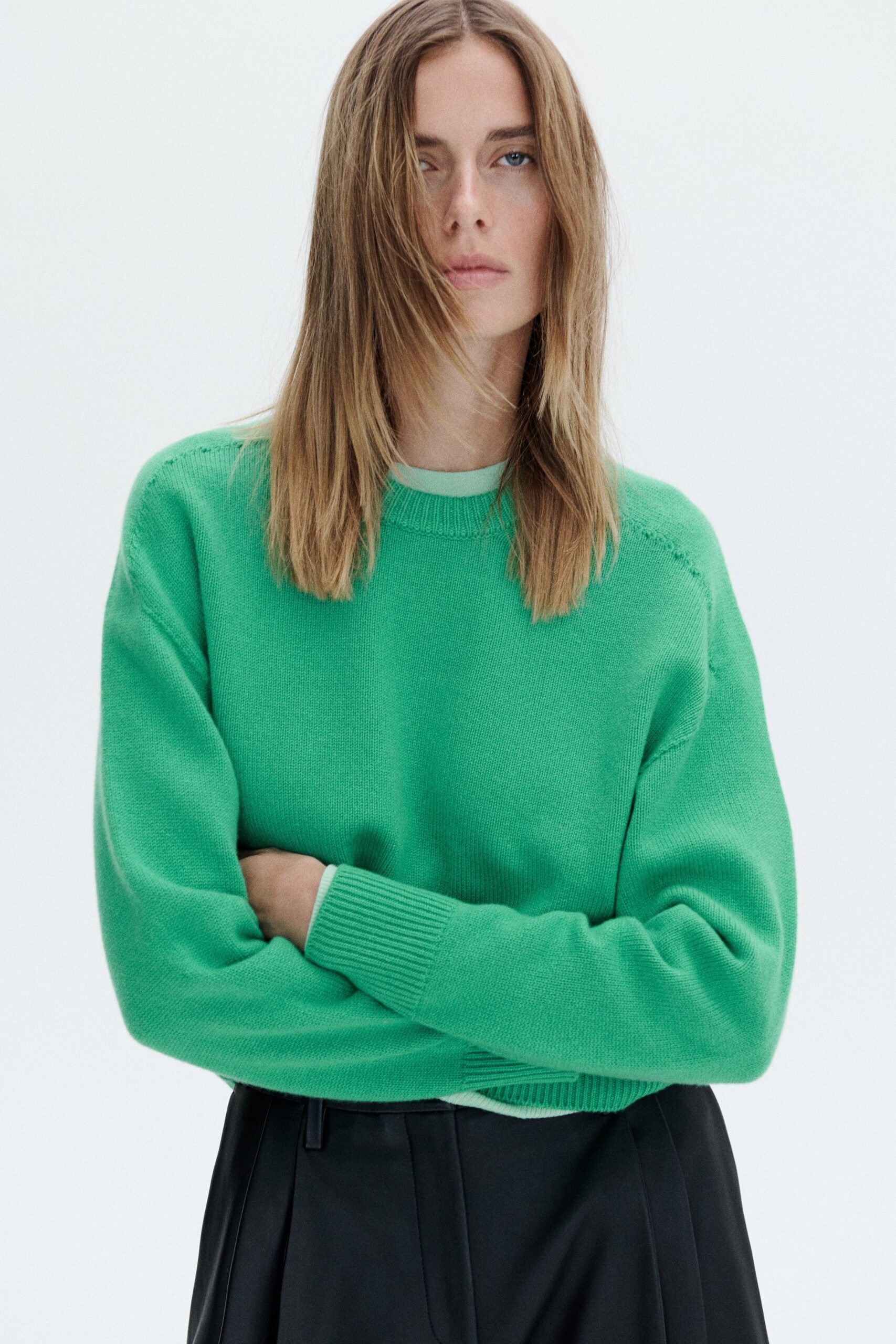 Mint crewneck sweater in fine gauge knit, pale tee edge peeking out. Black tailored trousers with deep front pleats falling from the waistband. First cool morning after a heatwave, office windows still cracked open.