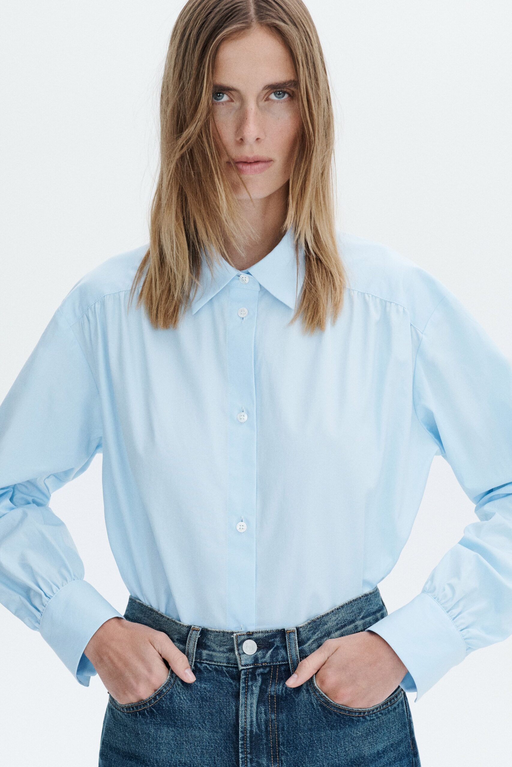 Powder-blue cotton shirt with a pointed collar and gathered cuff seams. Mid-wash denim jeans, front pockets opened by the hands. Office air-conditioning in August, lunch eaten at the desk.