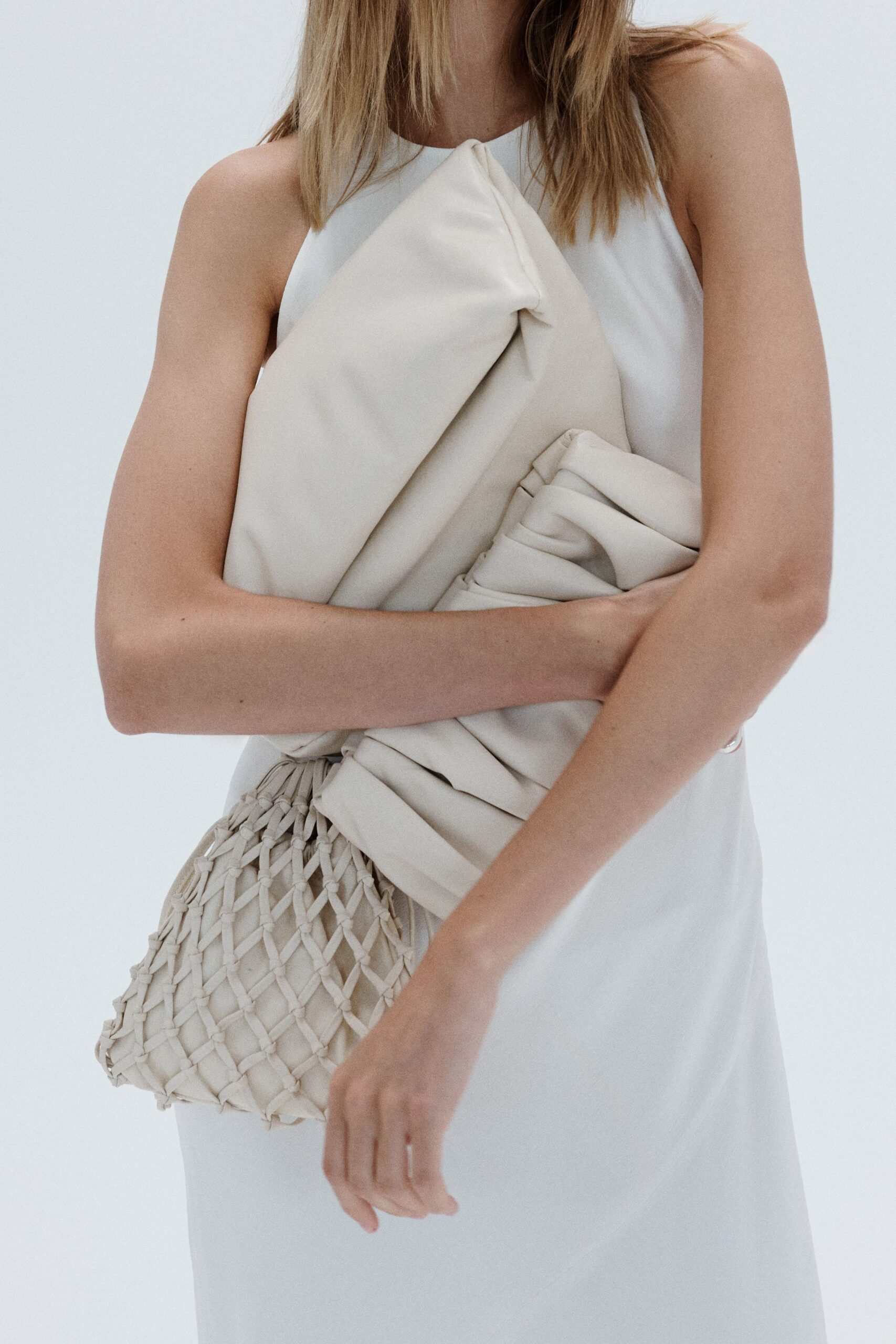 Chalk halter dress with a wide folded panel knotted at the throat. Soft pouch bag under a hand-knotted lattice sleeve. For carrying flowers home after a civil ceremony at noon.