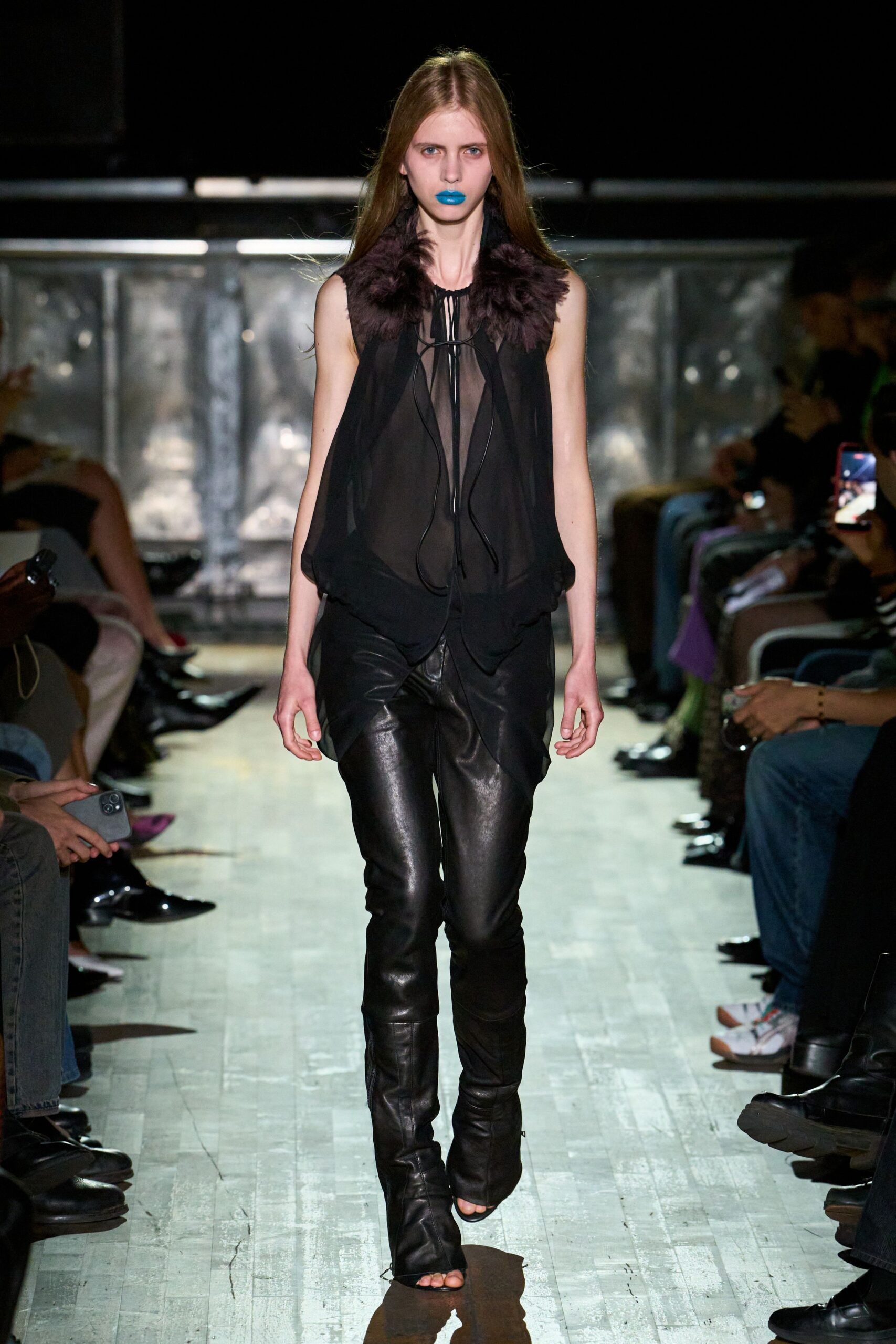 Sheer black chiffon top with a smoky feather collar and dangling ties. Black leather trousers cut close, creased bright along the thighs. Open-toe leather boots slouching slightly at the ankle. Walking under strip lights after midnight, lipstick still louder than your voice.