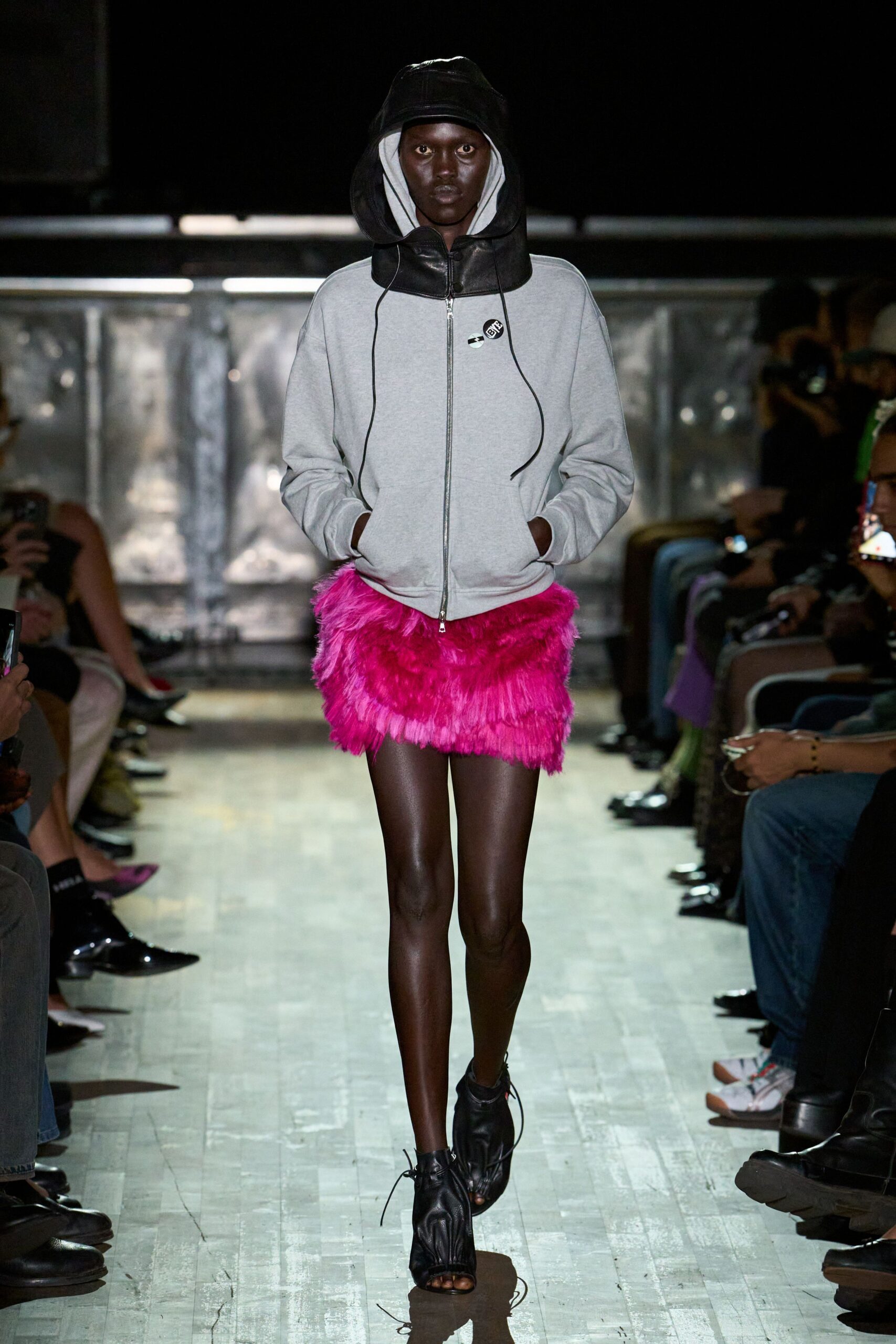 Pale grey zip hoodie under an oversized black leather hood, cords hanging long. Hot-pink shaggy faux-fur mini, layered in rough horizontal swishes. Black peep-toe ankle boots, laces trailing loose across the floor. Walking the club corridor after coat check, neon already staining the carpet.