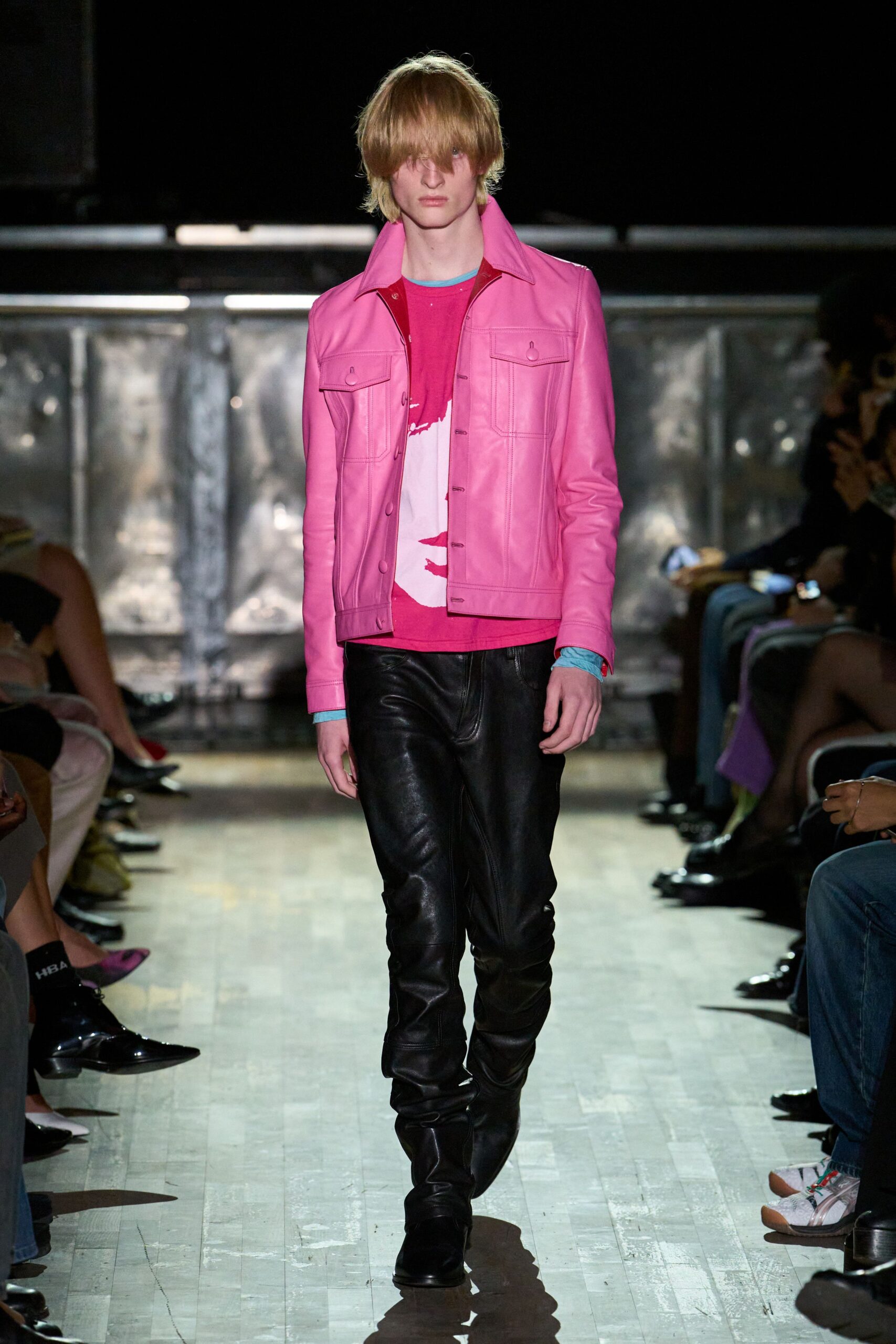 Bubblegum leather trucker jacket with flap pockets and a high spread collar. Hot-pink graphic tee over pale aqua jersey cuffs. Black leather trousers, stacked hard into the ankle. Sharp black boots disappearing under the hem. Walking to the corner shop after bleaching your hair in a friend's bathroom.