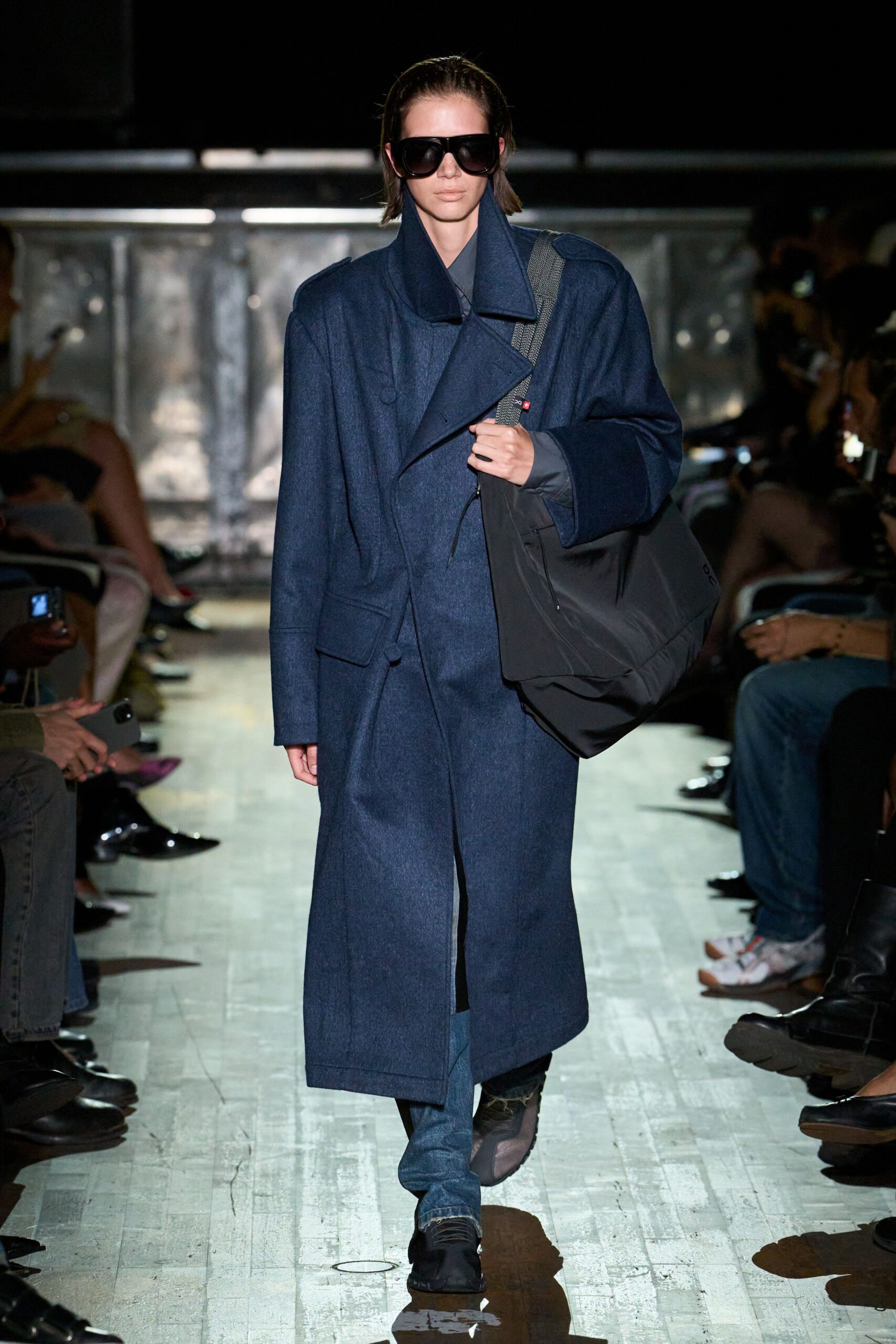 Oversized black shield sunglasses. Ink-blue wool overcoat, doubled lapels wrapped high at the throat. Slate technical shirt peeking from the cuff. Slouchy black tote on a grey webbing strap with red tab. Blue jeans dragging into frayed hems. Black split-toe slip-ons. Walking into the station before dawn with one bag too many.