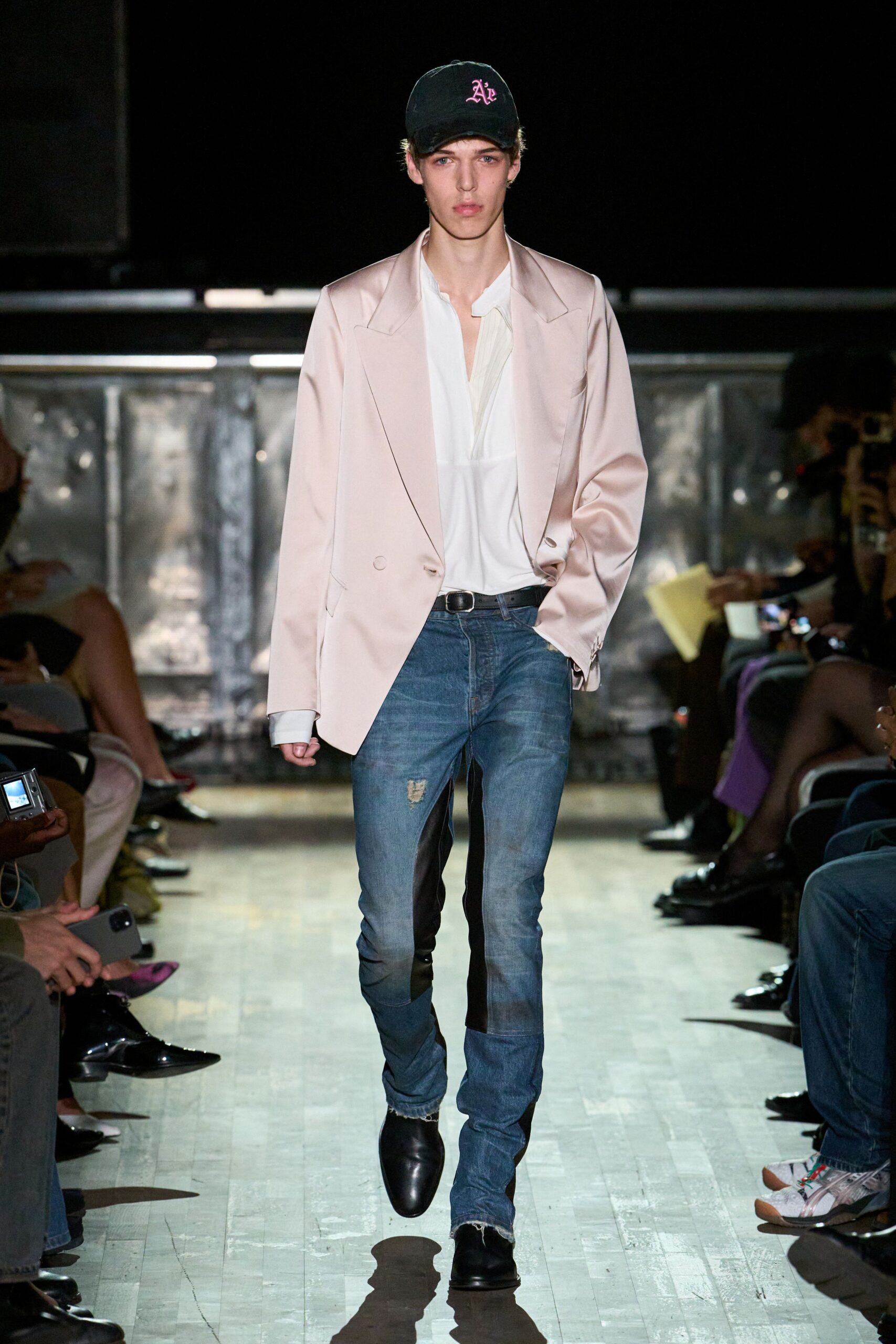 Black cap with pink Gothic initials. Blush satin blazer, shoulders broad and slightly dropped. White blouse with a deep split placket and soft gathers. Faded blue jeans, one knee nicked and hems fraying. Black square-toe boots. Walking into a small-town casino still wearing yesterday's dinner jacket.