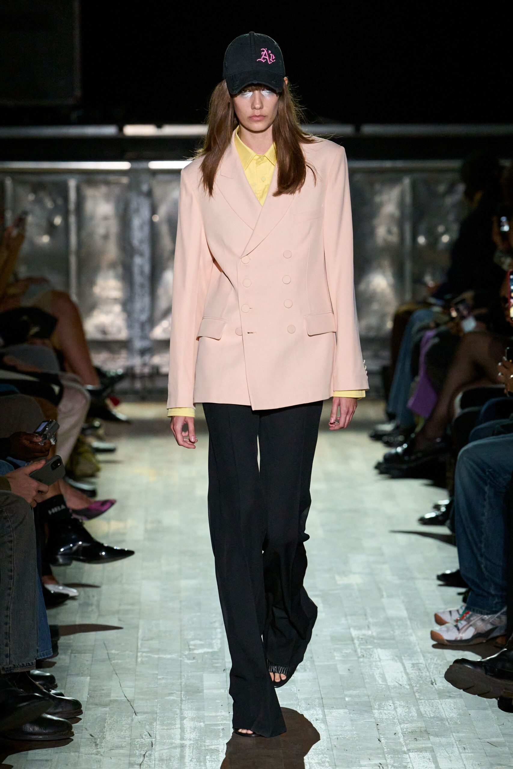 Black cap with pink gothic monogram embroidery. Pale lemon shirt buttoned to the collar. Blush double-breasted blazer, one button left hanging open. Black trousers pooling over the foot. Black open-toe heels. Walking into a municipal hearing after missing three hours of sleep.