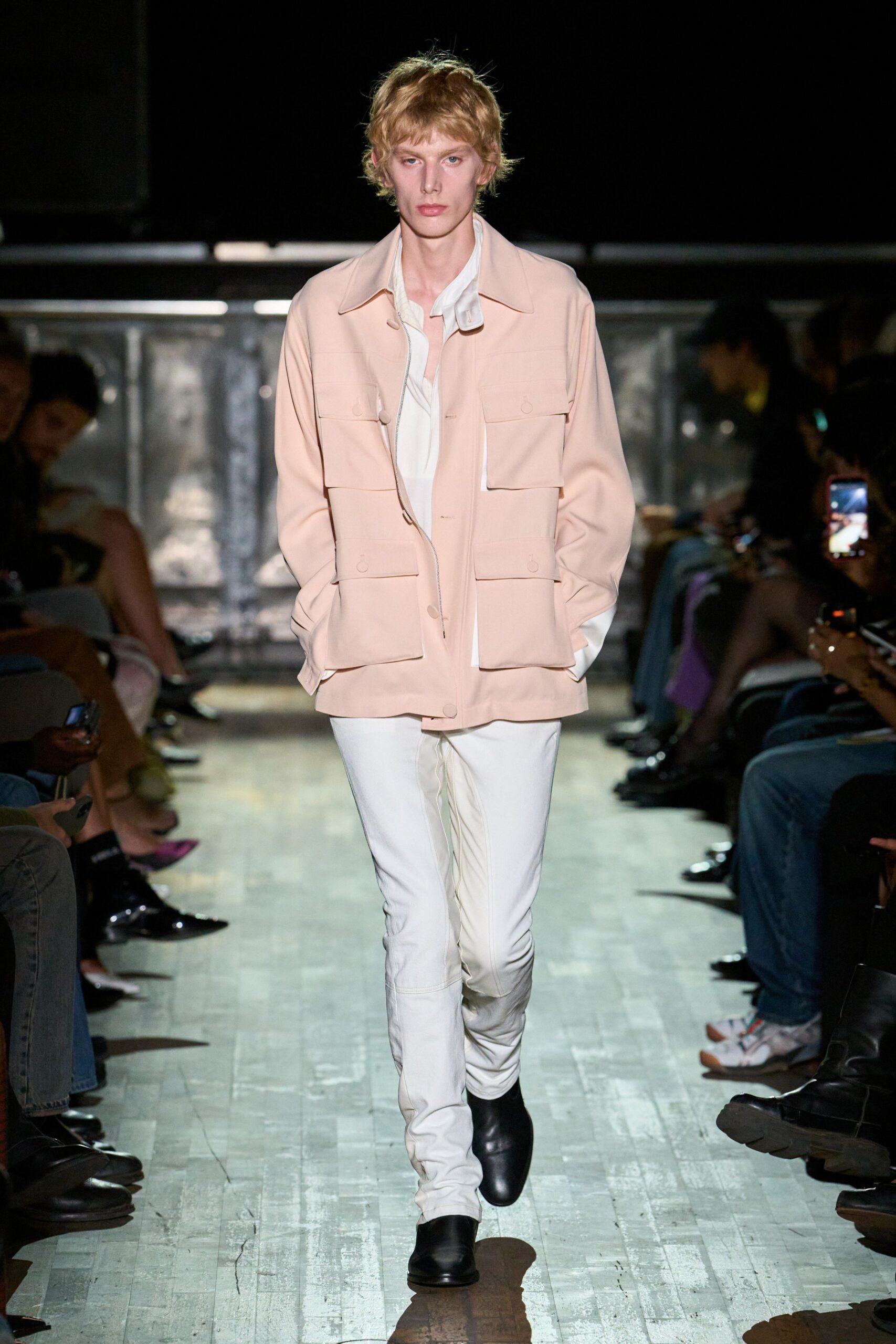 Blush utility jacket with four bellows pockets and a half-dropped zip pull. Ivory shirt hanging open at the throat, one collar point flipped up. White fitted leather trousers, creased hard through the knees. Black square-toe ankle boots. Walking into a hotel lobby before breakfast, still carrying last night's heat.