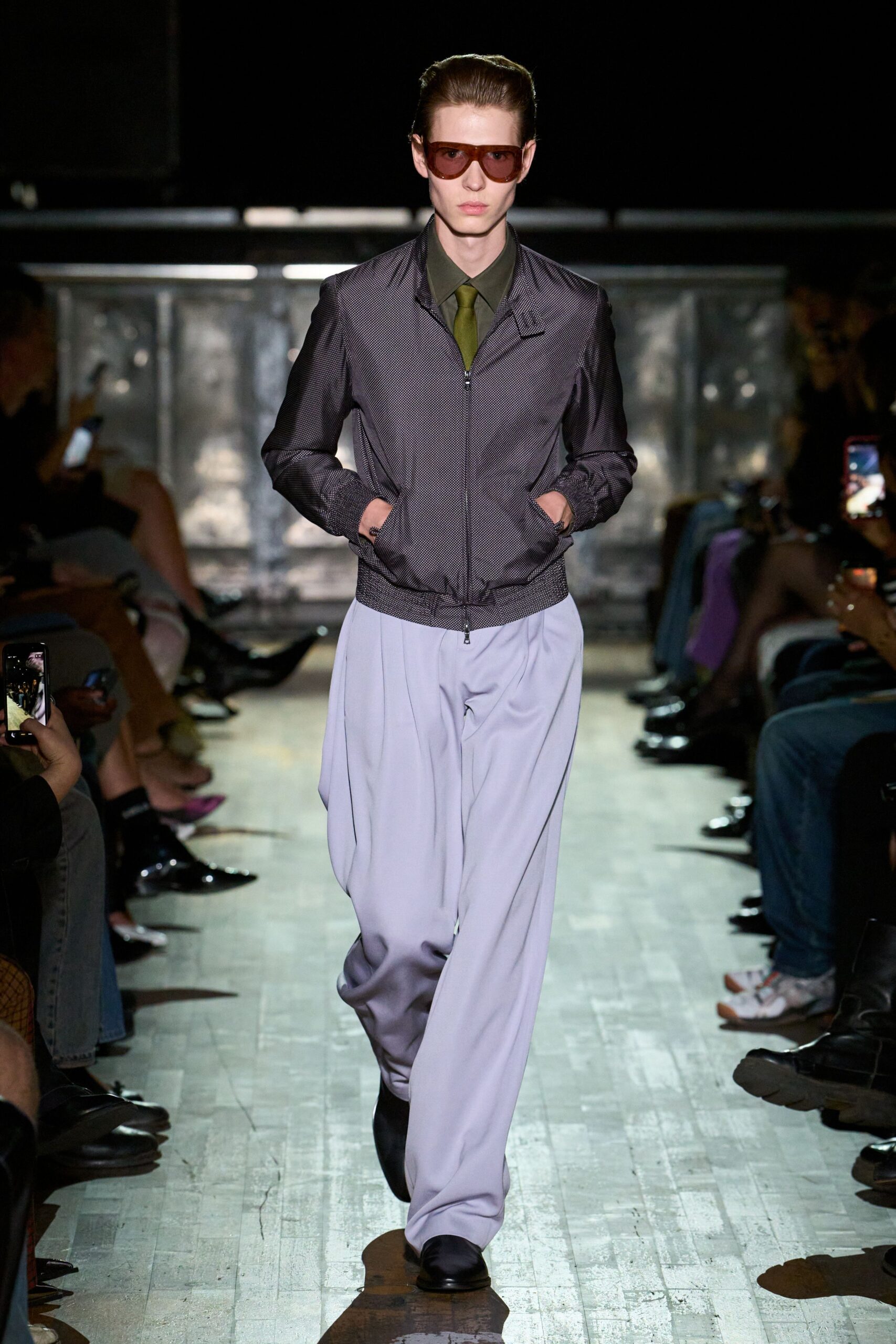 Amber wraparound visor shades. Pepper-flecked zip blouson with smocked cuffs and hem. Olive shirt buttoned tight under the collar. Moss satin tie, narrow and flat. Pale dove trousers cut wide with deep front pleats. Black square-toe shoes. Walking into a civic building after rain, tie still damp at the knot.