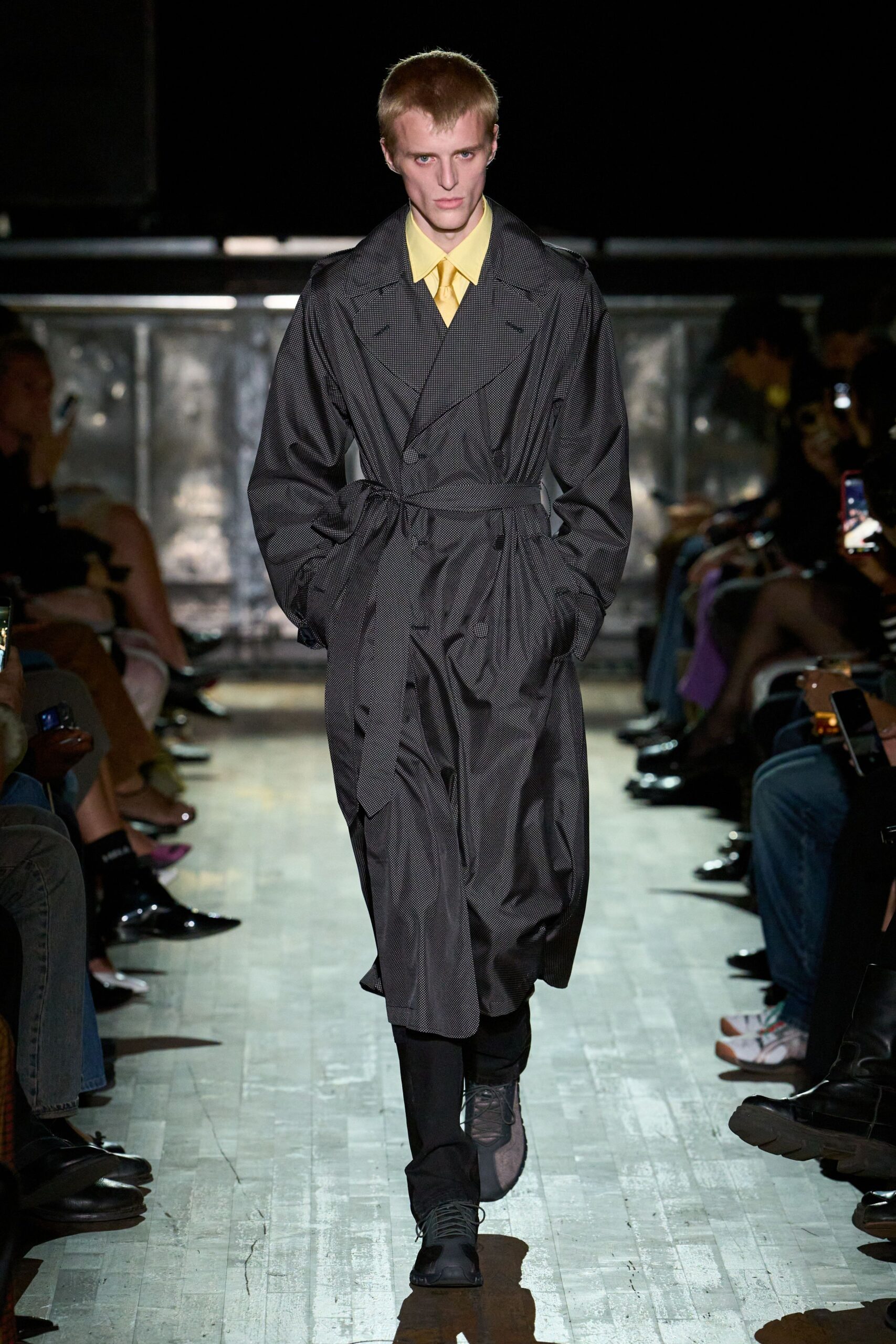 Butter-yellow shirt with a long pointed collar and matching tie. Charcoal micro-check trench, double-breasted and cinched with a drawcord waist. Black trousers breaking softly over the instep. Dark lace-up shoes with squared rubberised toes. Walking to a courthouse through drizzle, tie still too bright for the weather.
