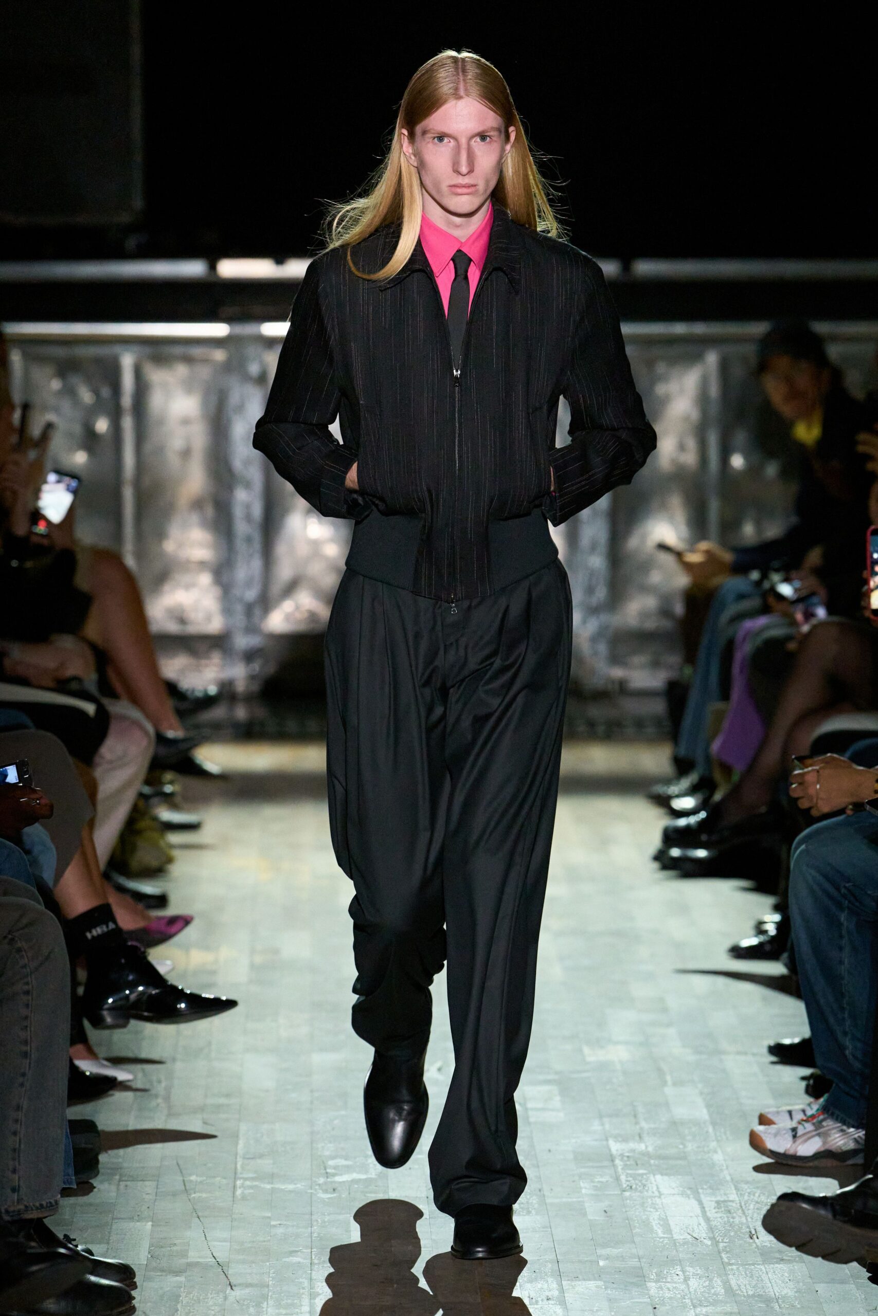 Hot-pink dress shirt buttoned tight beneath the tie knot. Narrow black tie, hanging straight and spare. Black pinstriped zip jacket with a broad ribbed waistband. Loose black trousers breaking long over the shoe. Square-toed black leather boots. Walking into a municipal wedding hall still smelling faintly of rain.