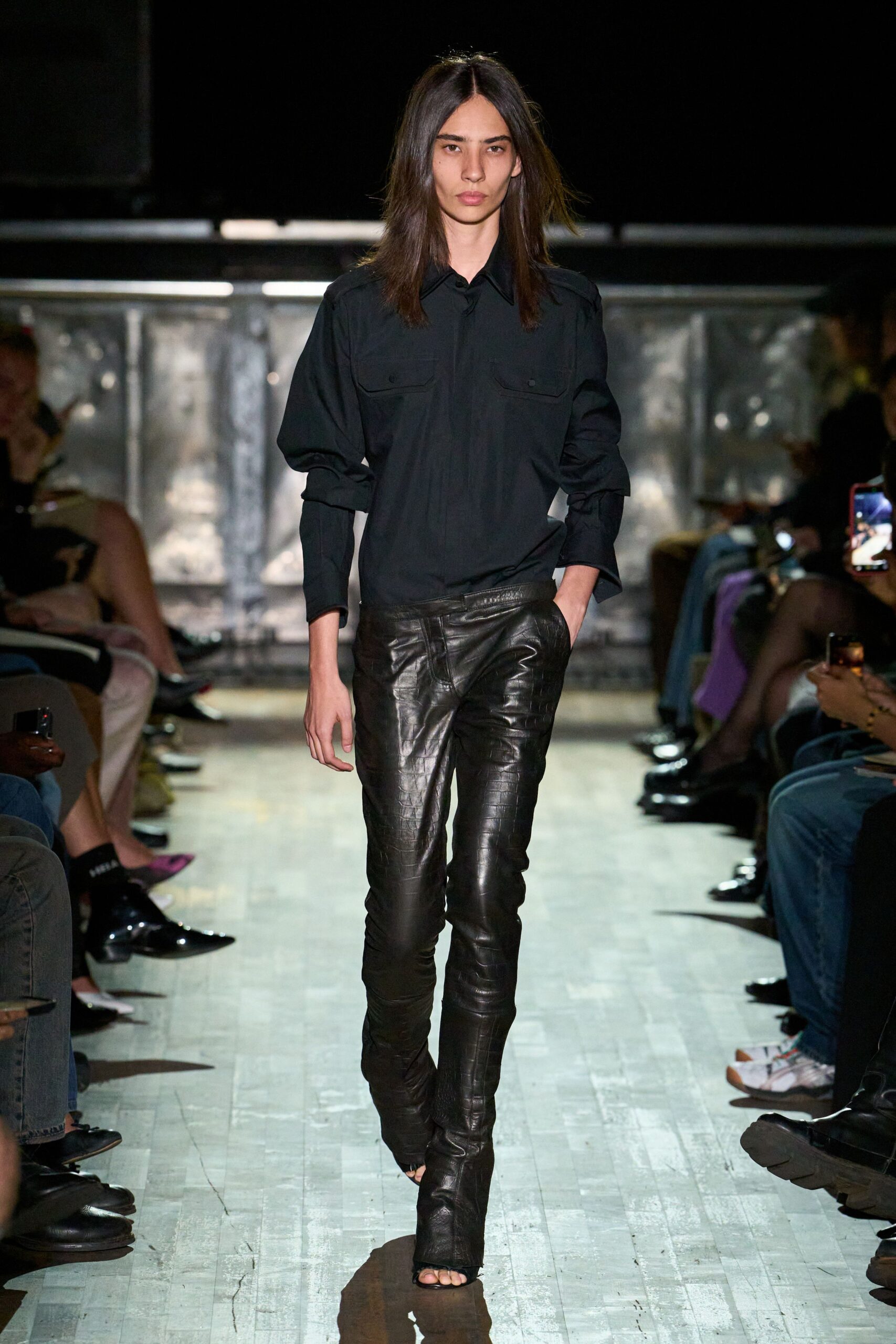 Black utility shirt with flap chest pockets and softly bunched sleeves. Black crocodile-embossed leather trousers, cut skin-tight through the leg. Open-toe leather boots with a slight break over the foot. Walking into a hotel bar before anyone has turned the lamps on.