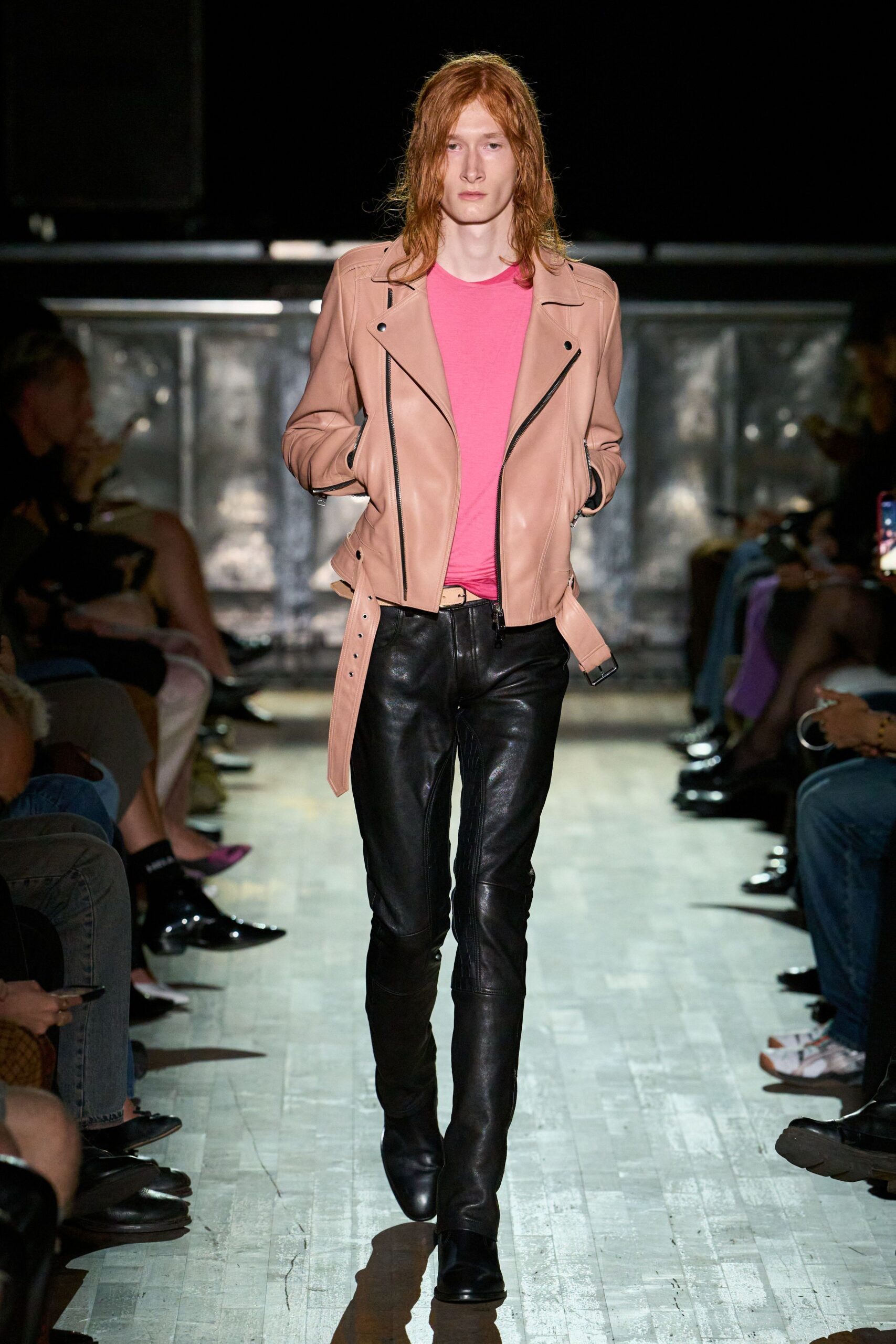 Blush leather biker jacket with black zips and one belt end hanging long. Hot-pink crewneck tee, softly slouched into the waistband. Black leather trousers with curved knee panels and a close shine. Square-toe black boots. Crossing the forecourt after midnight, takeaway receipt still in a pocket.