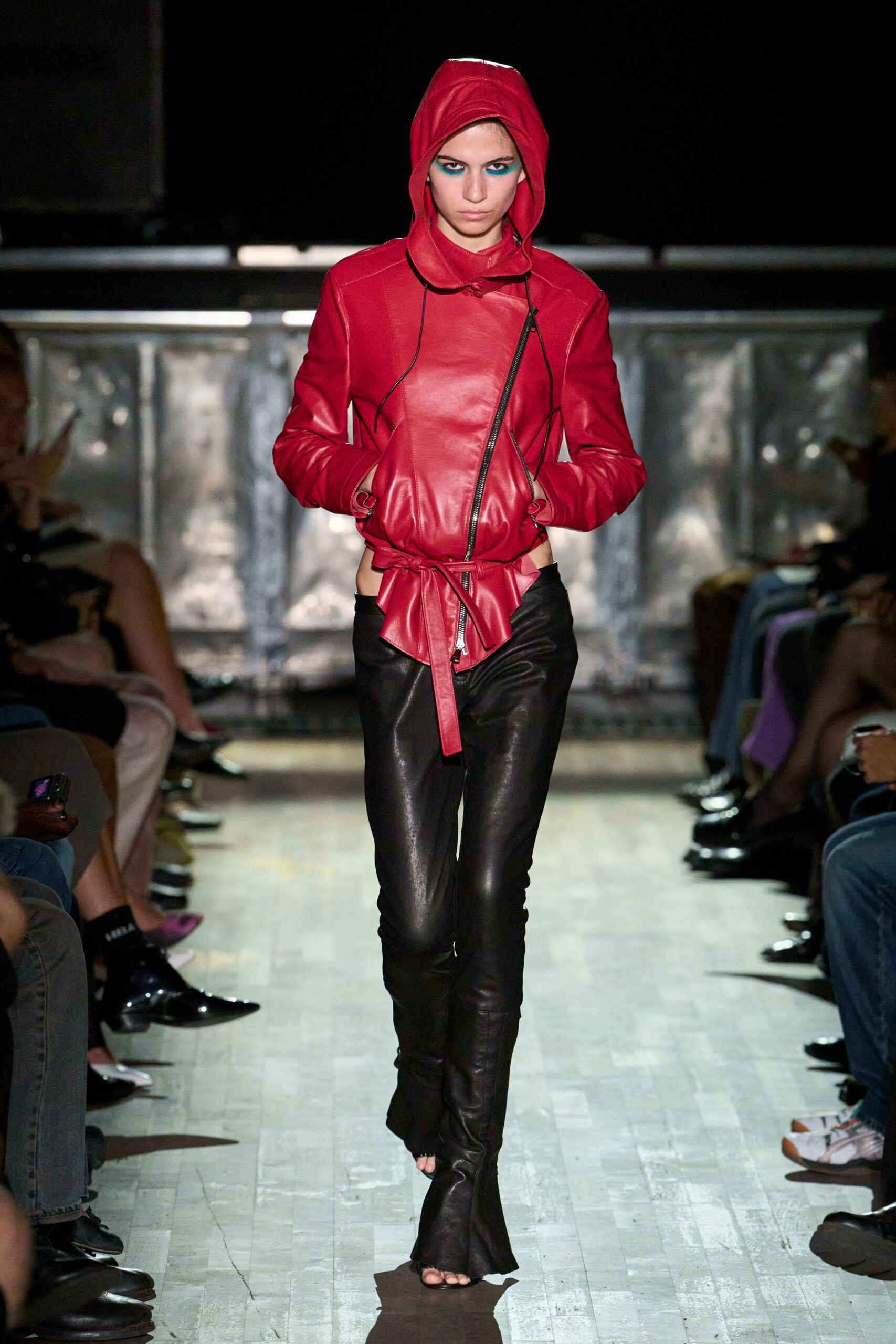 Tomato-red leather jacket with an off-center zip and scarfed hood. Long self-tie leather belt knotted low, one end hanging past the fly. Black leather pants cut skin-tight with split hems over the feet. Open-toe black leather boots peeking through the flared hems. Walking into a warehouse party before the heaters have kicked in.