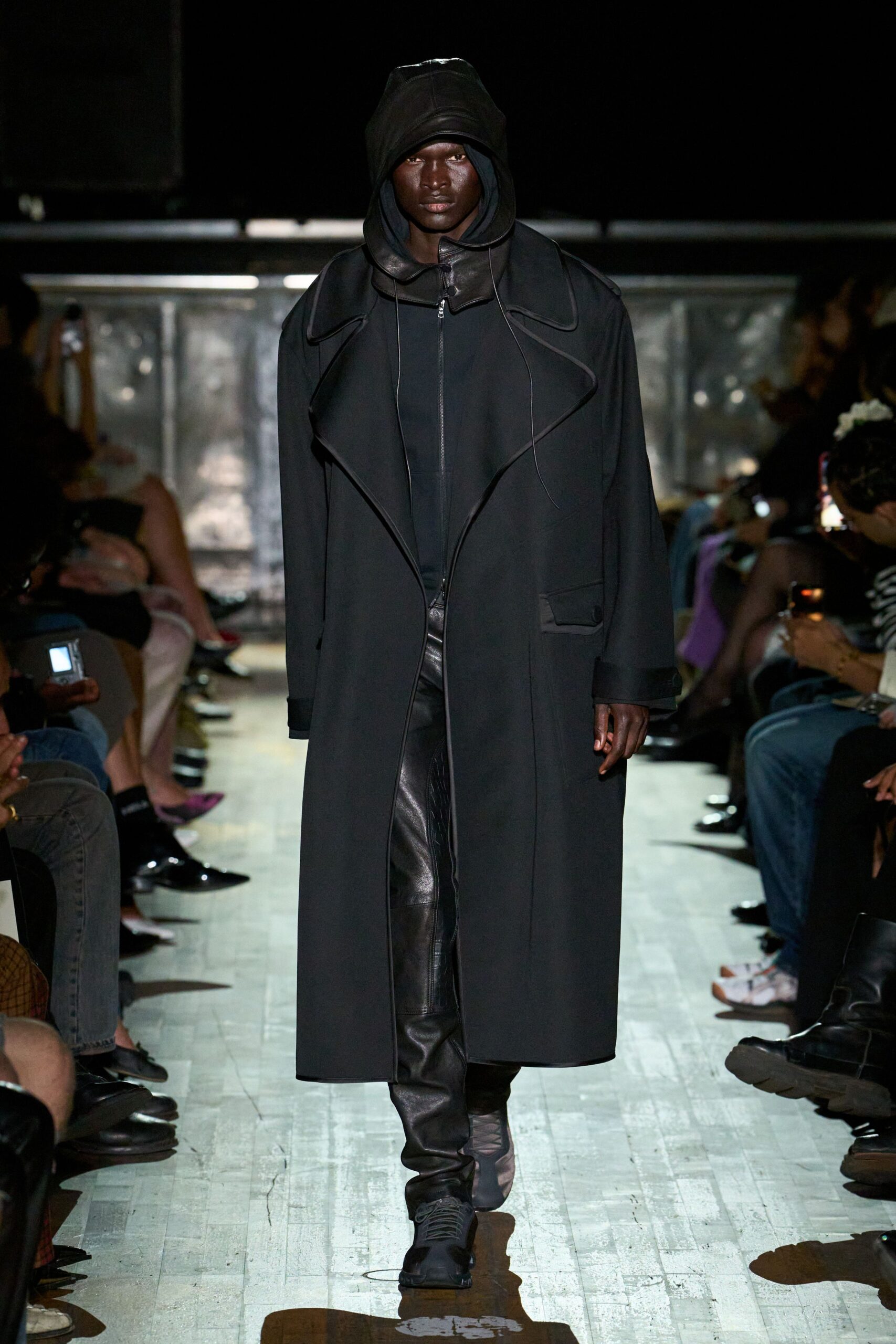 Black leather hood with a peaked crown and long hanging cords. Floor-length black coat, raw-edged lapels curling away from the zip. Black leather trousers, wrinkling hard through the thigh and shin. Square-toe black boots with crisscross lacing over the instep. Walking the river path after rain, keys cold in your pocket.