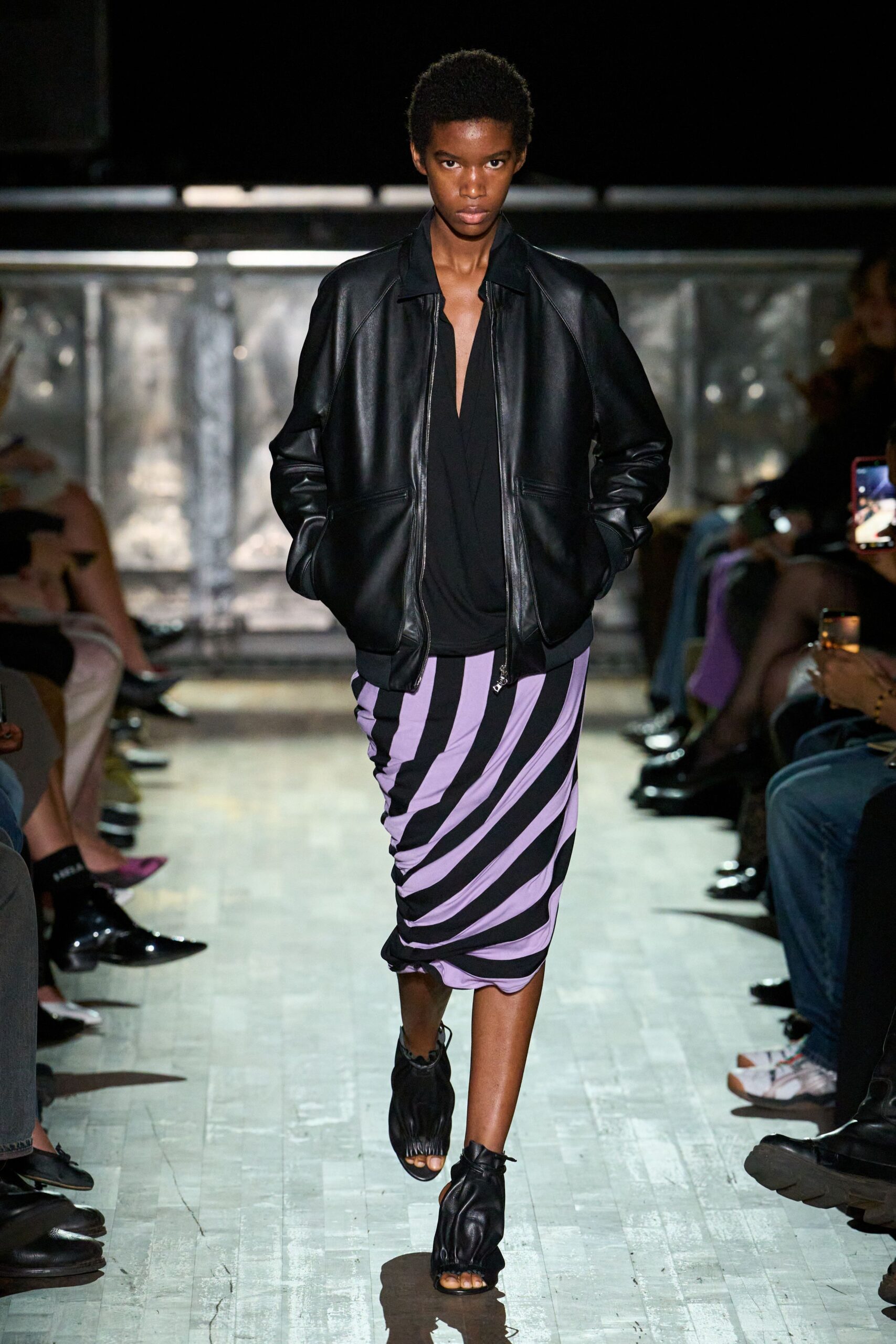 Black leather bomber with raglan shoulders and a dangling zip pull. Black draped jersey top cut to a narrow plunging V. Lilac and black jersey skirt, twisted into diagonal stripes. Black leather sandals, ruched over the foot with open toes. Walking the hotel corridor for ice at dawn, still dressed from yesterday.