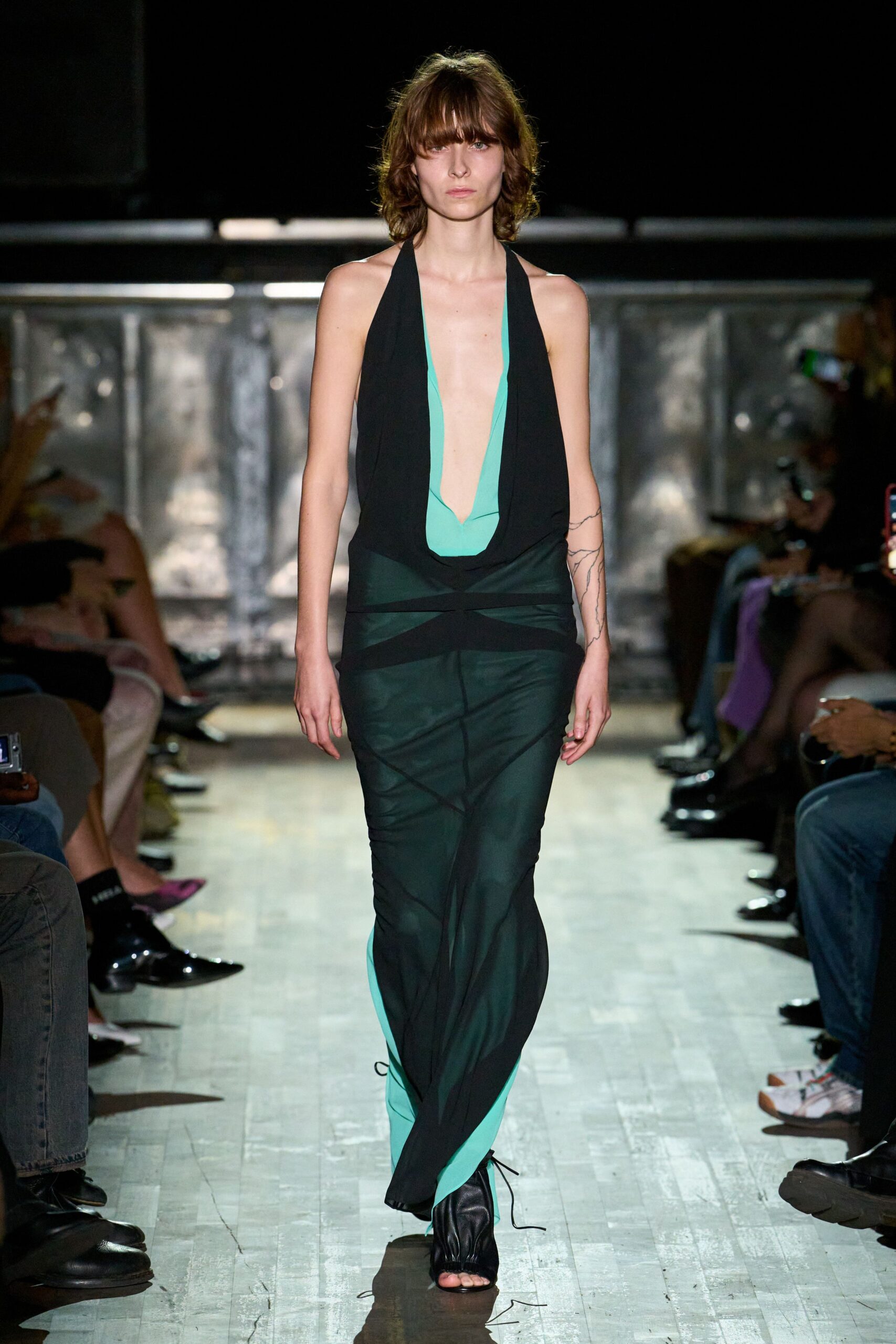 Black halter gown with a seafoam plunge and sheer wrapped skirt panels. Black gathered leather sandals, thin ties trailing loose on the floor. Crossing a hotel ballroom after midnight with the air-conditioning still too cold.