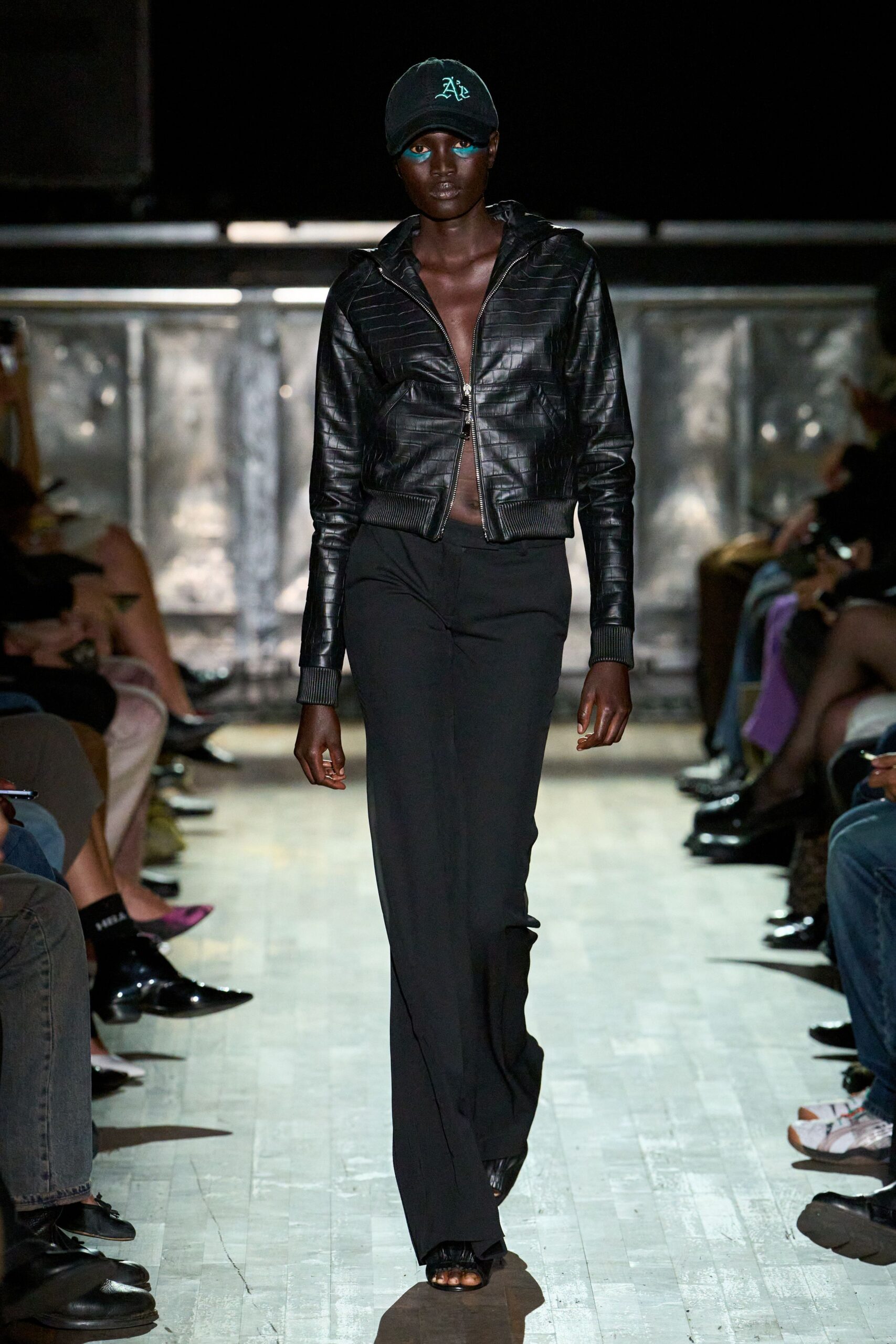 Black baseball cap with pale stitched lettering across the crown. Black crocodile-embossed zip hoodie, ribbed cuffs and hem hugging tight. Floor-grazing black trousers, soft drape breaking over the front shoe. Black open-toe heels with a gathered cuff at the ankle. Walking from a basement club into the wet shine after rain.