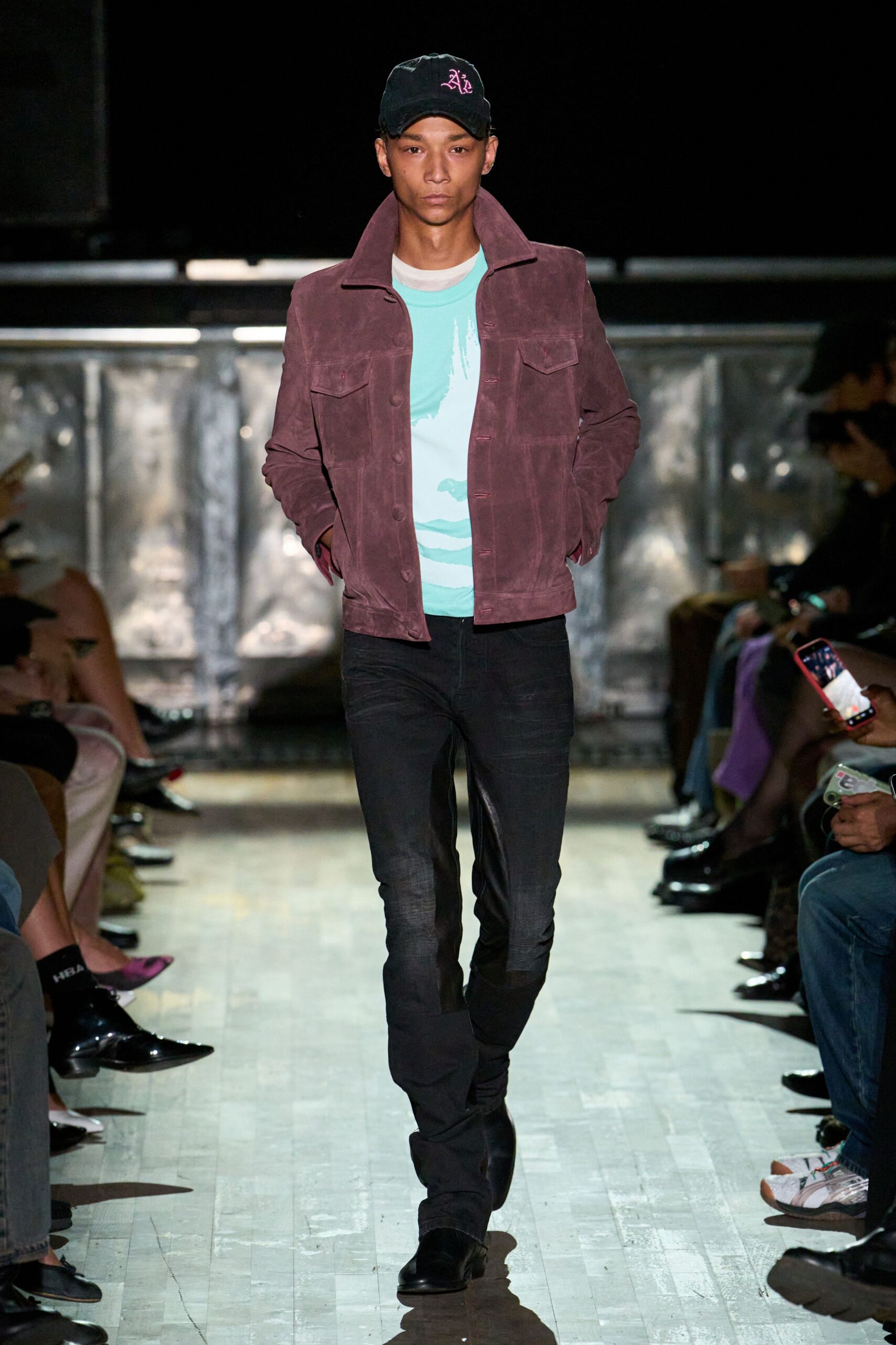 Black baseball cap with a pink Old English monogram. Plum suede trucker jacket, point collar flipped high and covered buttons. Aqua tee washed with a cloudy white brushstroke print. Black skinny trousers, fading dusty at the thighs. Squared black boots. Walking the boardwalk after rain, amusement lights not switched on yet.