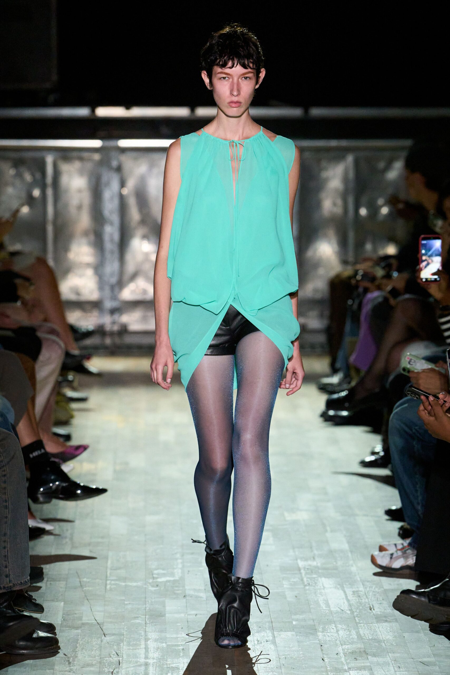 Mint chiffon tunic, slit neckline tied with a narrow cord loop. Black leather micro shorts peeking beneath the draped split hem. Icy lilac shimmer tights. Black leather peep-toe ankle boots, laces trailing loose on the floor. Crossing a hotel ballroom after midnight with heels in your hand.