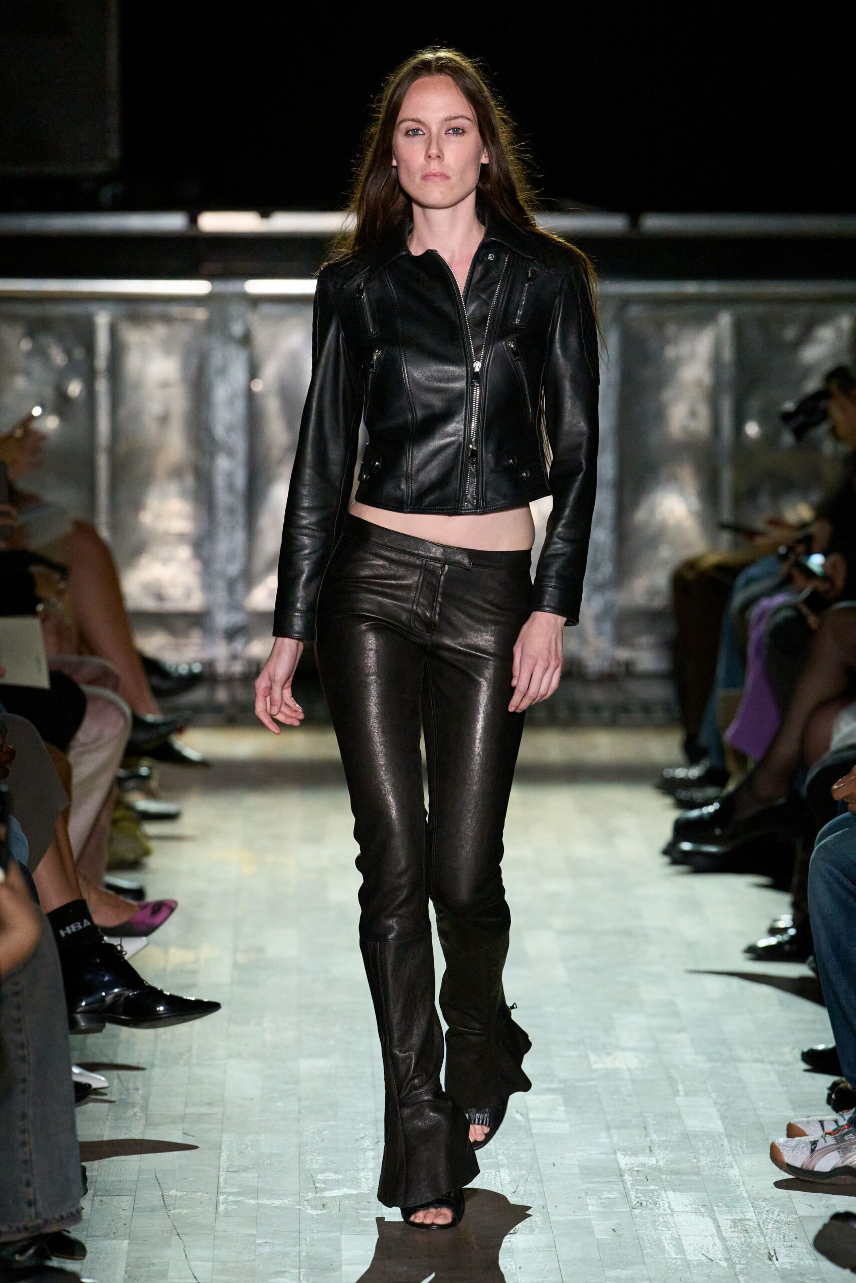 Black leather moto jacket, zipped chest pockets and a cropped curved hem. Low-rise black leather flares, whiskered at the hips and pooling slightly. Black open-toe heeled sandals. Walking out of a basement club into wet pavement and first light.