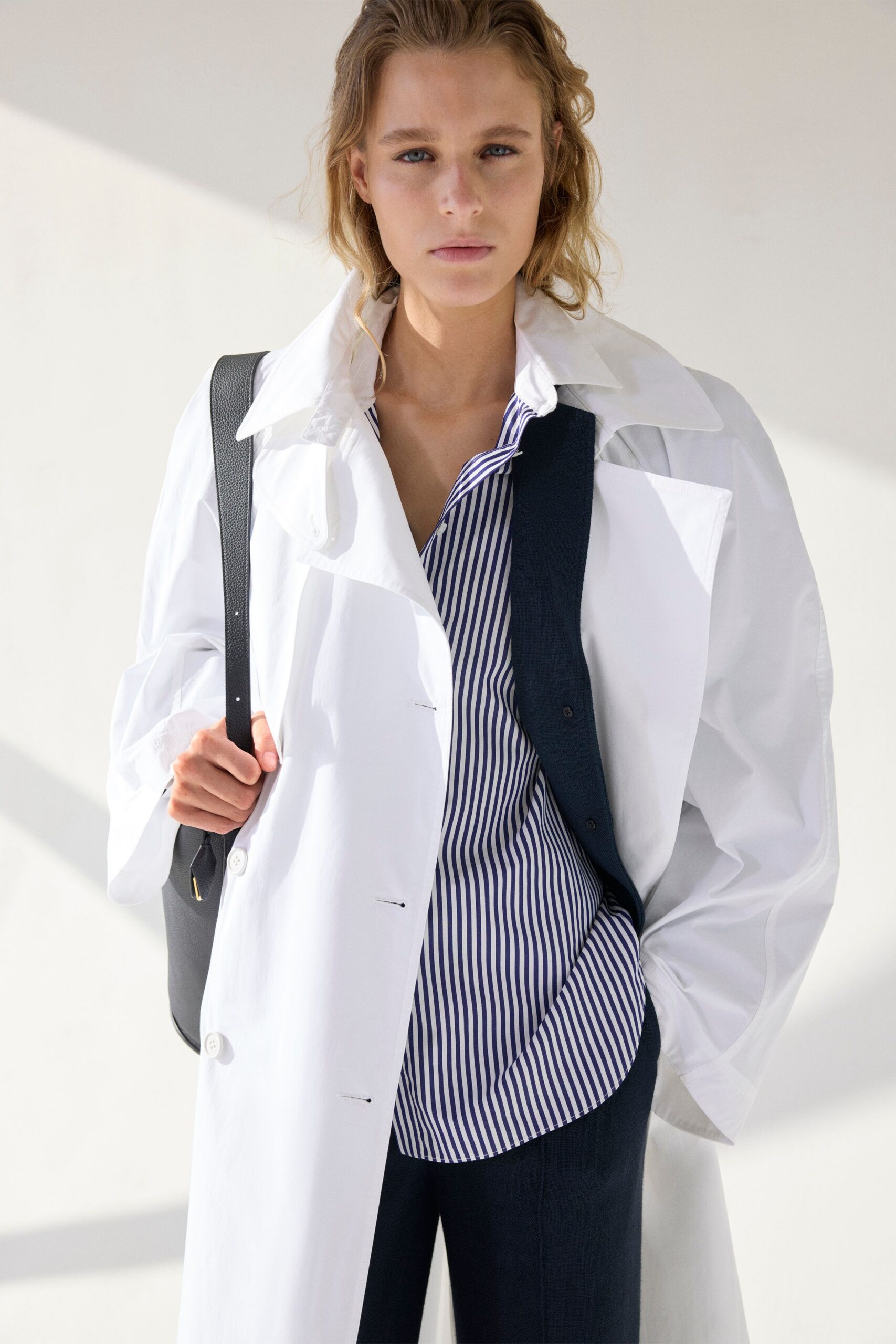 White cotton coat with a blown-up collar and pin-fastened placket. Blue-and-white banker-stripe shirt, collar slipping open under the lapel. Dark tailored trousers with a pressed front crease. Black pebbled leather, thin strap gripped in one fist. Early train to the office after sleeping somewhere else.