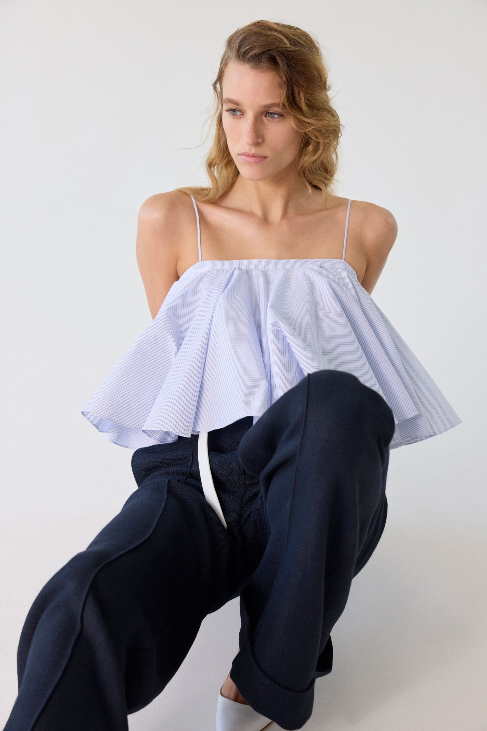 Pale blue pinstriped camisole cut into a wide sculpted peplum flare. Black wide-leg trousers in dry crepe, a white tie dropping at center. White leather mules. Morning light in an apartment still echoing after the movers left.