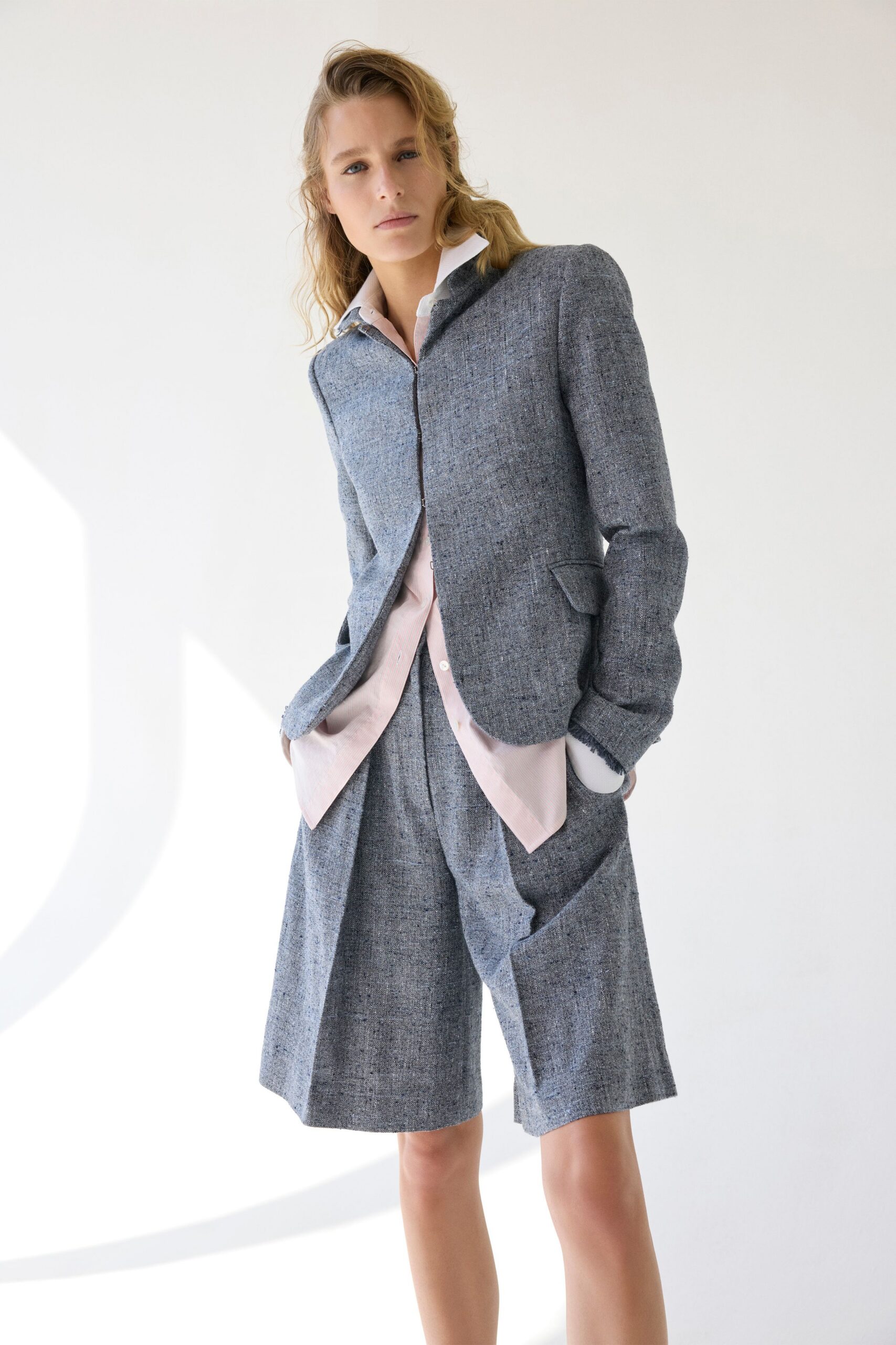 Pale blush shirt with a white collar and long pointed hem. Salt-and-pepper tweed jacket, hook-fastened, with frayed cuffs turned back. Matching tweed bermuda shorts cut wide through the leg. Morning meeting in a glass office before the air conditioning settles.