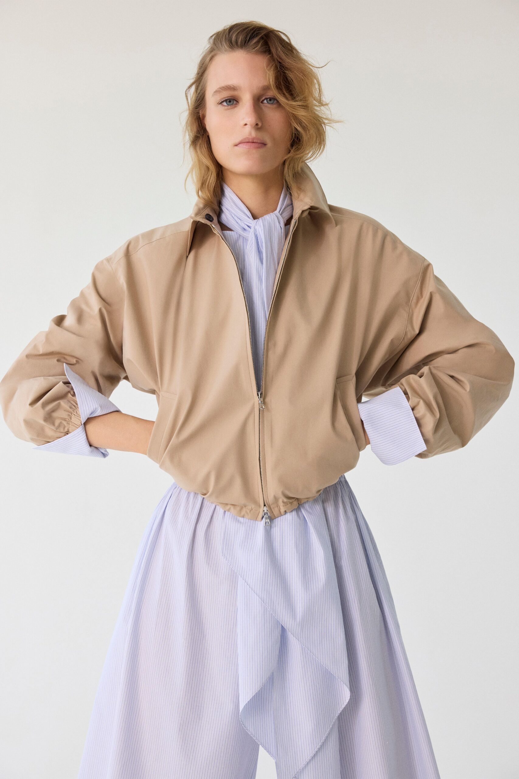 Sand cotton blouson with a standing collar and two-way zip. Pale blue pinstriped dress, scarf neck knotted close and one ruffle falling diagonally. Walking into the office early enough to hear the radiators start.